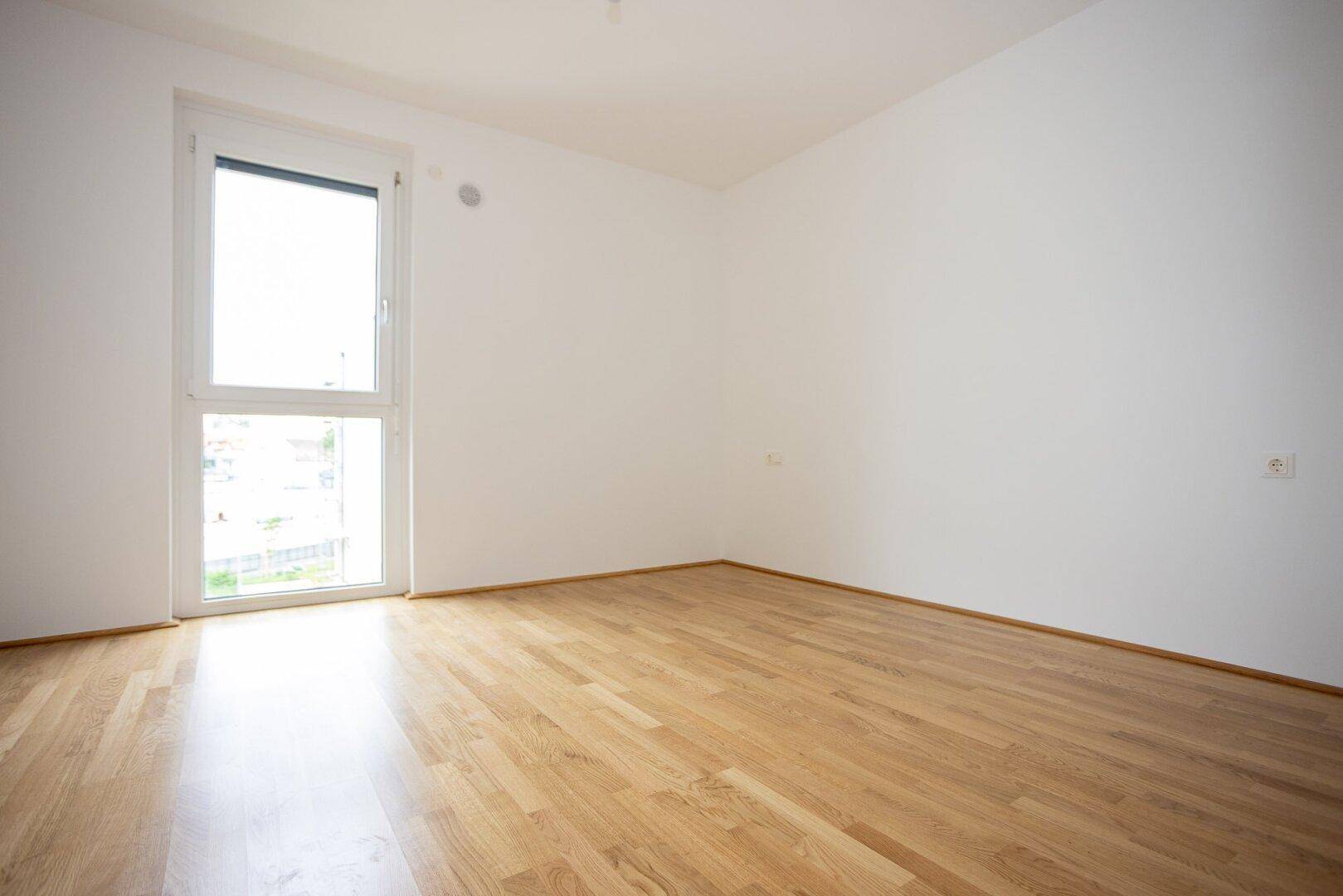 Helles, unmöbliertes Schlafzimmer mit Parkettboden und großem Fenster, ideal für viel Tageslicht.