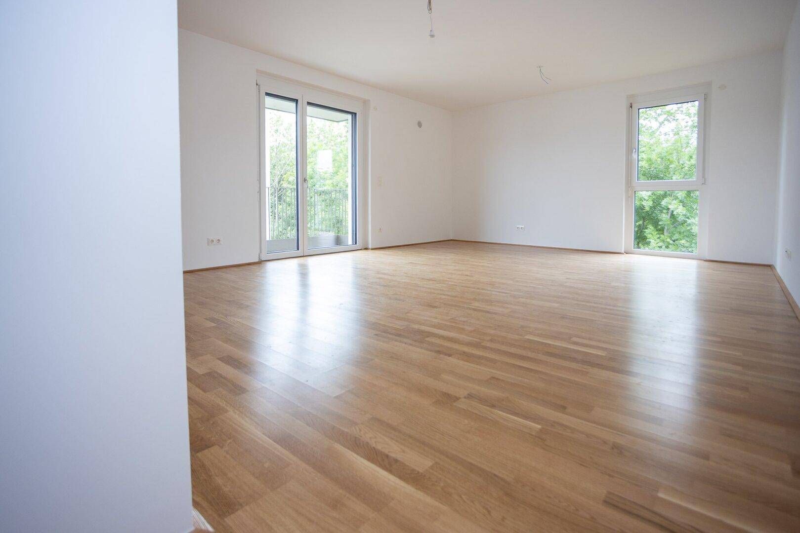 Helles, leeres Wohnzimmer mit Parkettboden und Zugang zum Balkon.