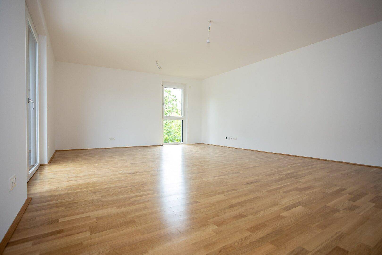 Lichtdurchflutetes, leeres Zimmer mit Parkettboden und großem Fenster.