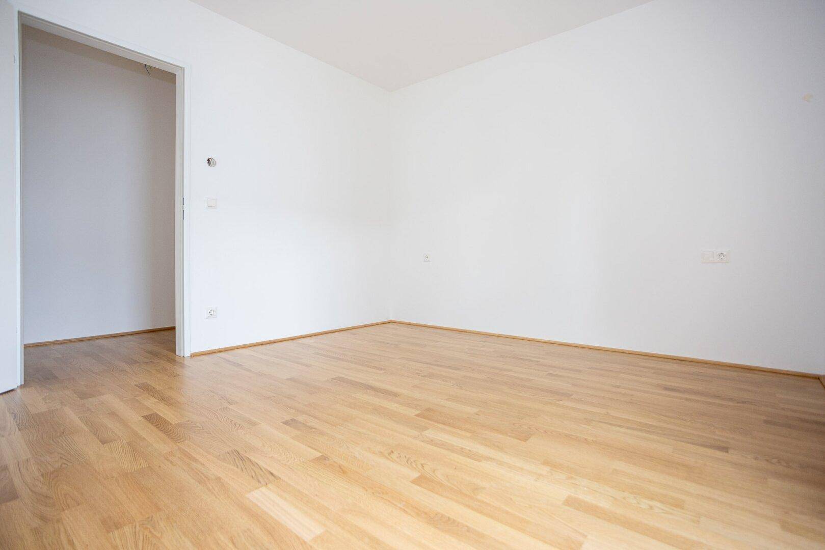 Unmöbliertes Zimmer mit hellem Parkettboden und weiß gestrichenen Wänden.