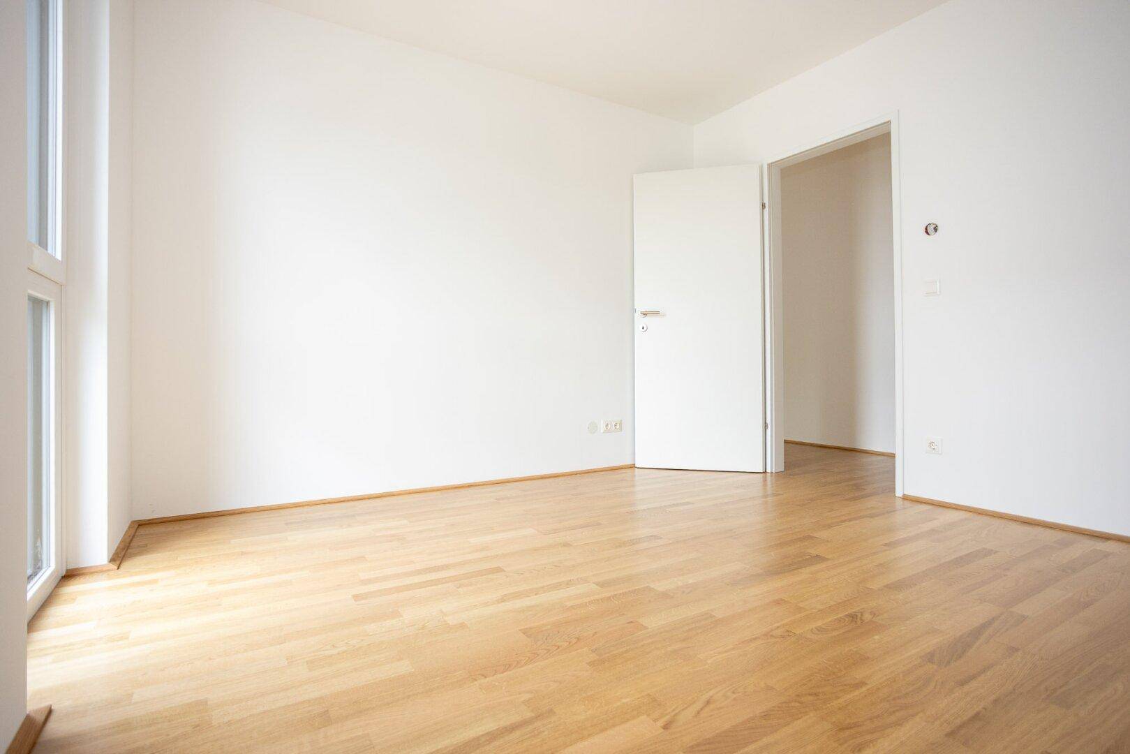 Helles, unmöbliertes Zimmer mit Parkettboden und Zugang zu einem weiteren Raum.