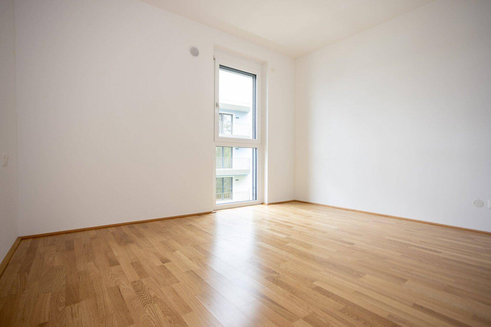 Leeres Zimmer mit hellem Parkettboden und hohem Fenster, ideal für Schlaf- oder Arbeitszimmer.