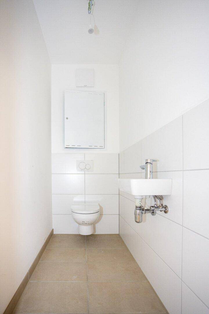 Funktionale Gästetoilette mit Wand-WC, kleinem Waschbecken und hellen Fliesen.