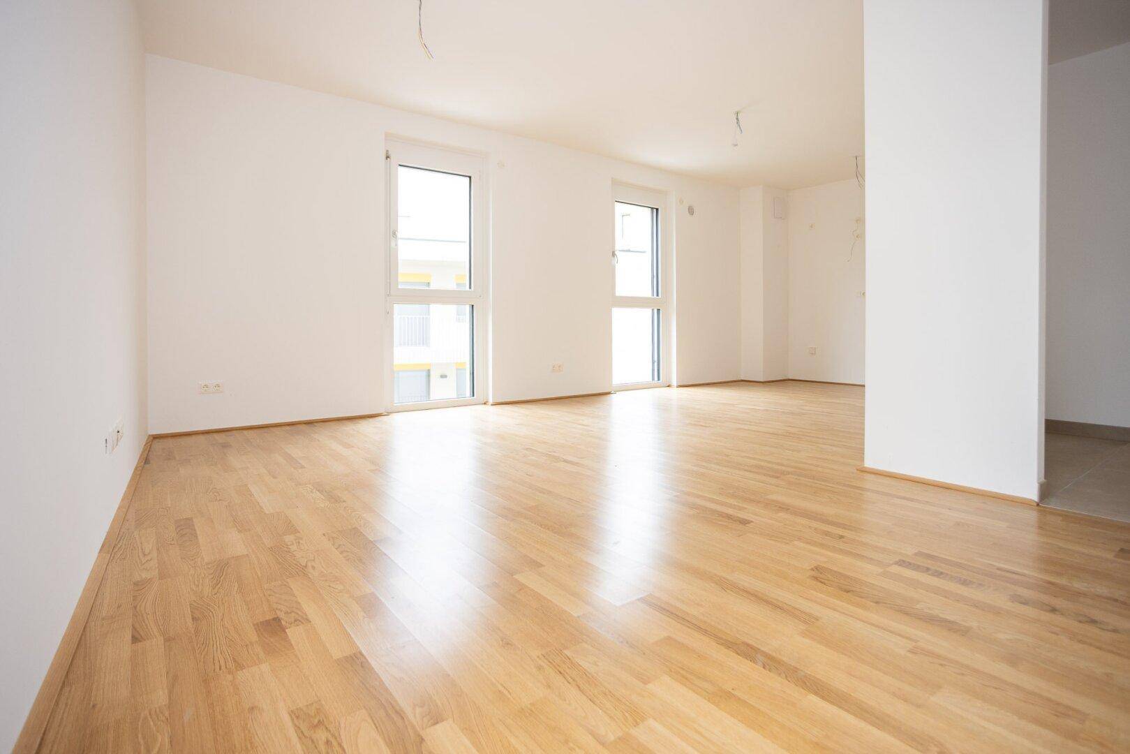 Heller, leerer Raum mit Laminatboden und zwei Fenstern, die viel Tageslicht hereinlassen.