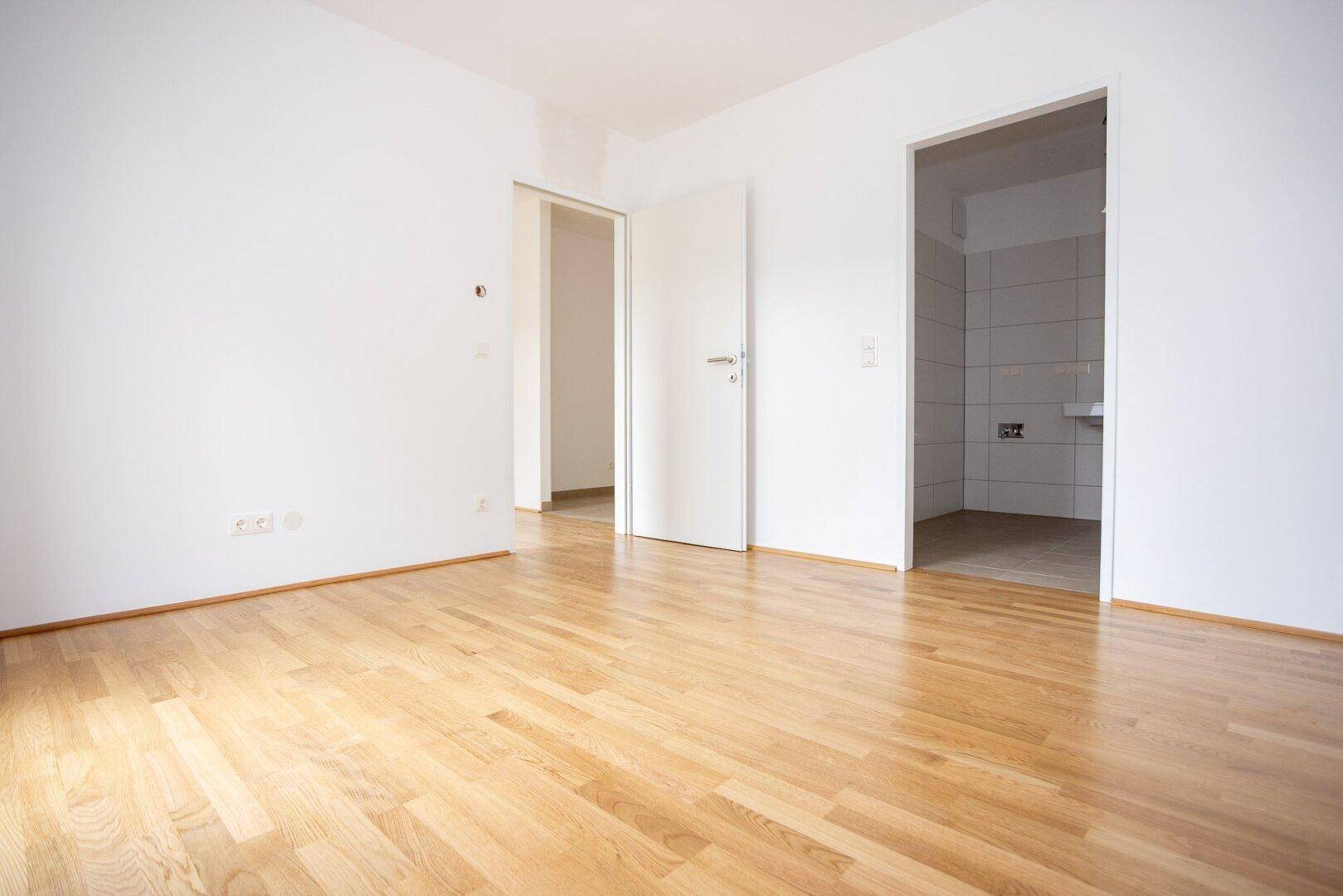 Leeres Zimmer mit Laminatboden und Zugang zu einem gefliesten Bereich, bereit zur Einrichtung.
