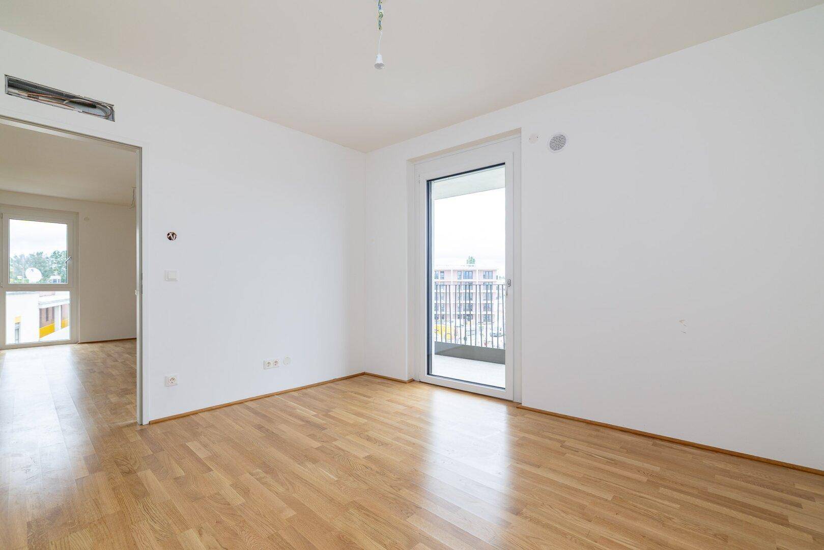 Leeres Zimmer mit Parkettboden und Balkontür, das viel Tageslicht empfängt.