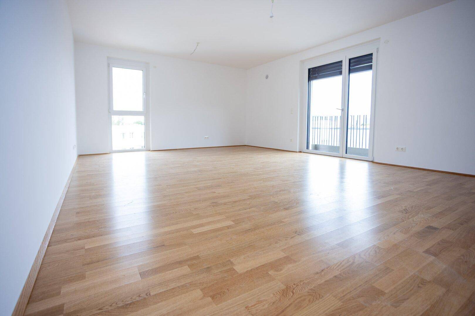 Helles, leeres Wohnzimmer mit glänzendem Parkettboden und Zugang zum Balkon.