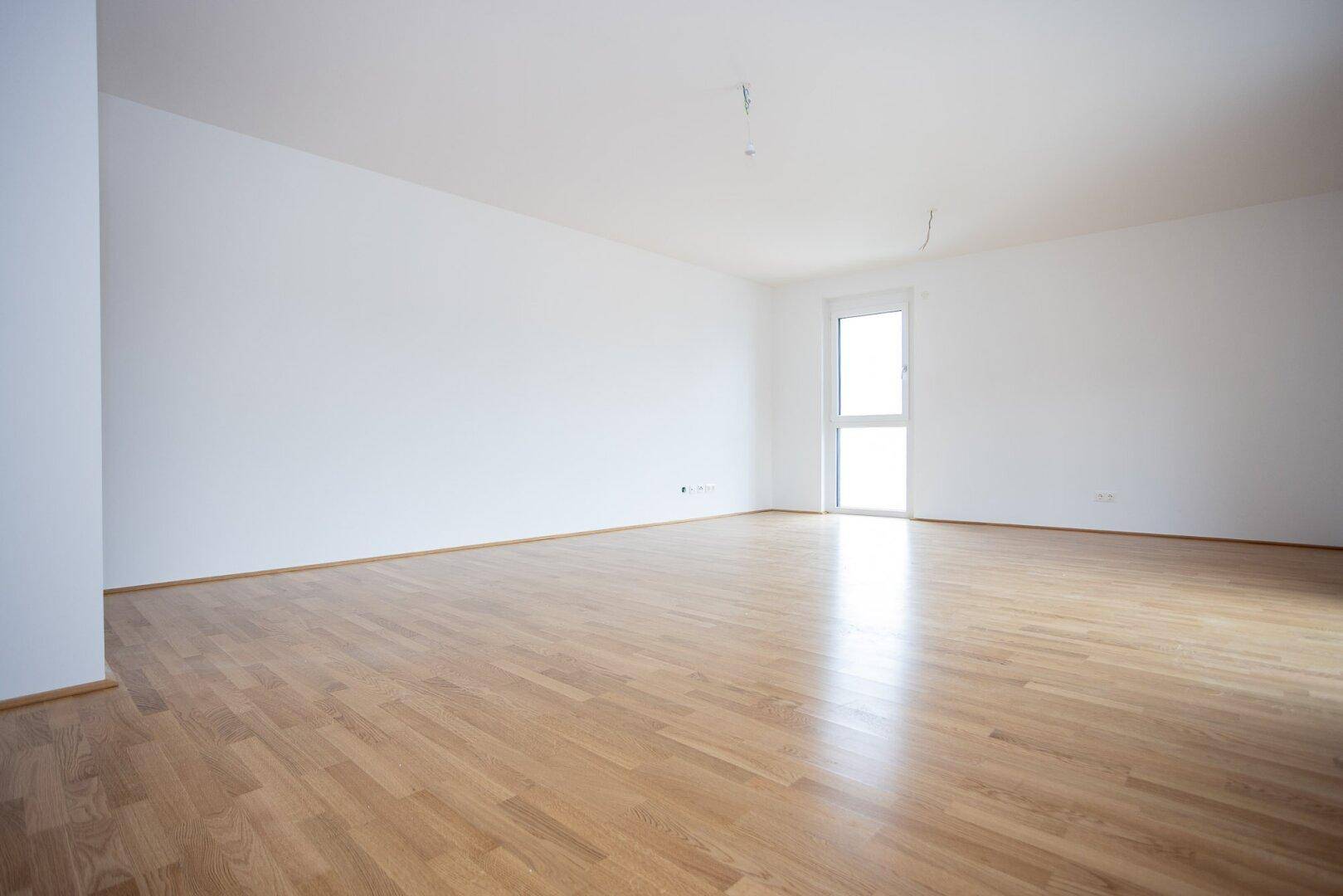 Geräumiges, helles Zimmer mit Holzboden und großem Fenster, bereit zur Einrichtung.