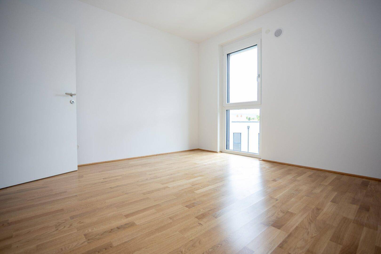 Leeres Schlafzimmer mit Holzboden und viel Tageslicht durch das hohe Fenster.
