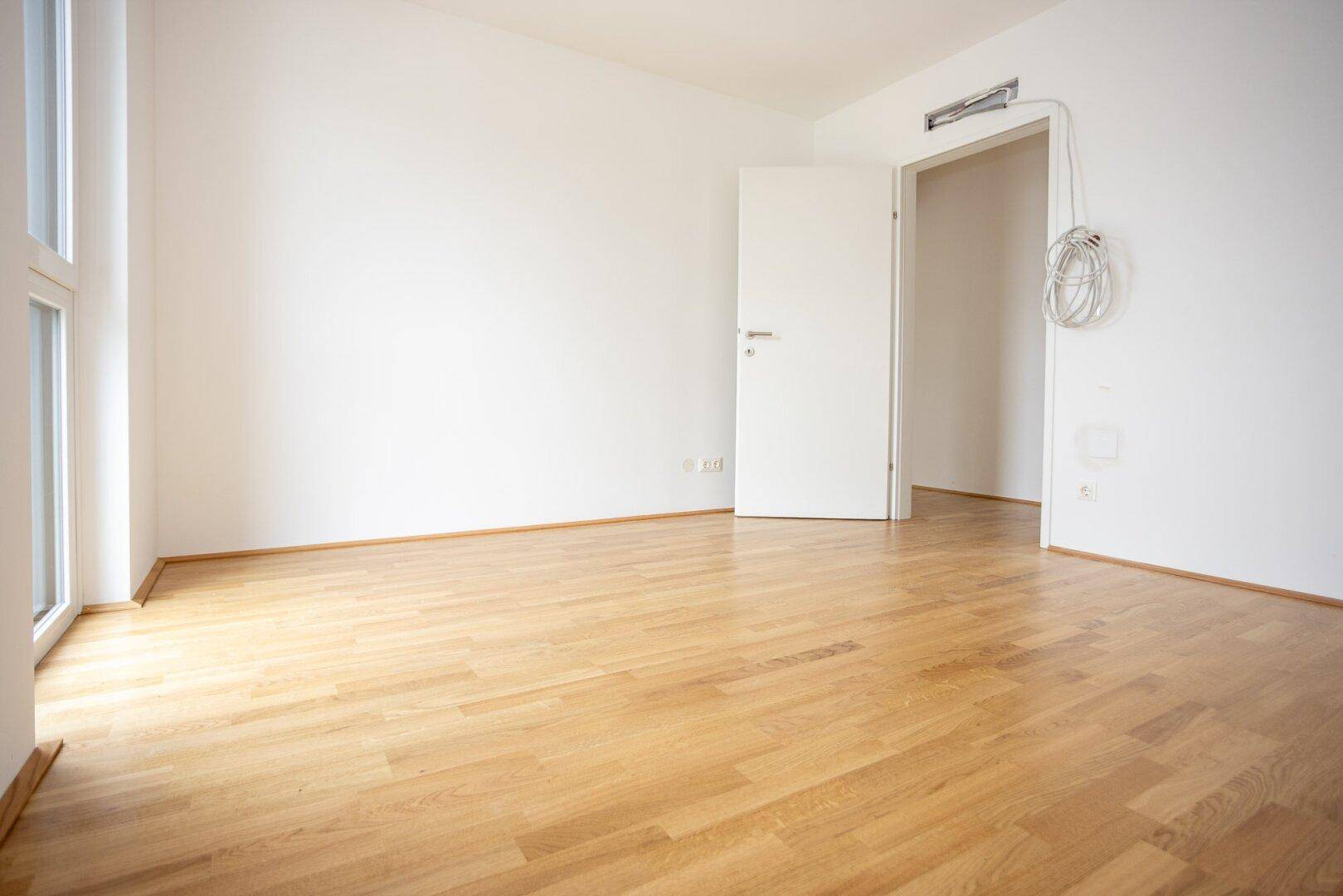 Leerer Raum mit Parkettboden und offener Tür zu einem weiteren Bereich der Wohnung.