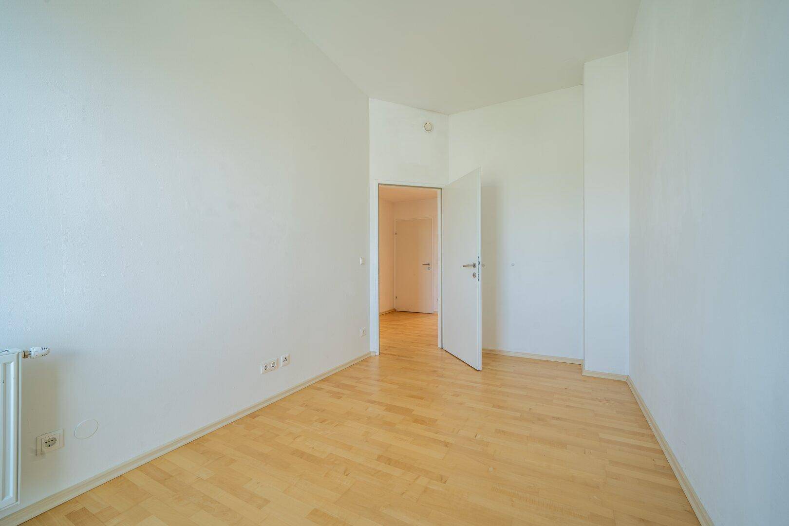 Leeres Schlafzimmer mit hellem Parkettboden und einer offenen Tür zu einem weiteren Raum.