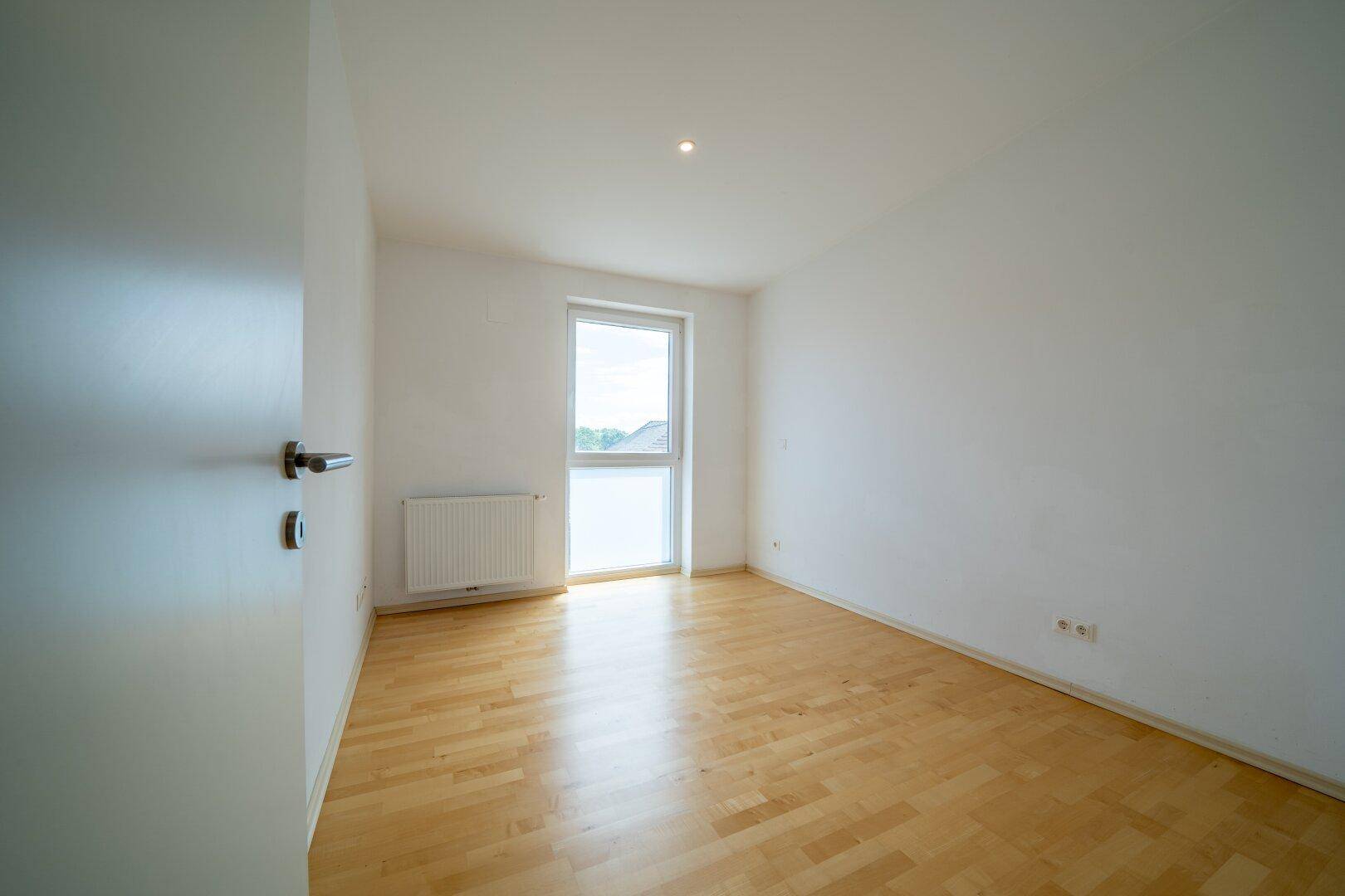 Schlafzimmer mit hellem Parkettboden und Fenster, das für natürliche Belichtung sorgt.