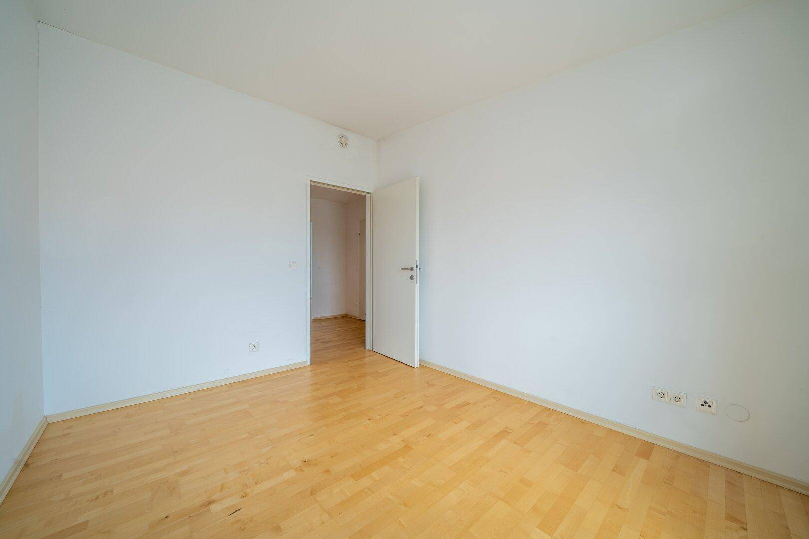 Leeres Zimmer mit Parkettboden und offener Tür, die den Zugang zum Flur zeigt.