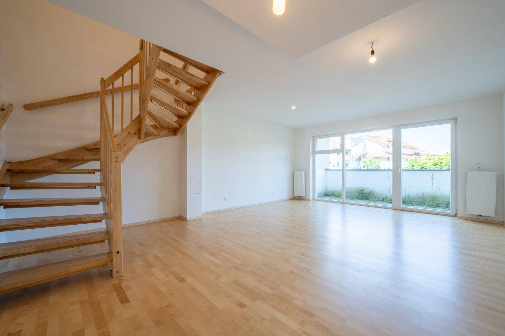 Geräumiges Wohnzimmer mit Holzboden und großen Fenstern, die viel Tageslicht hereinlassen.
