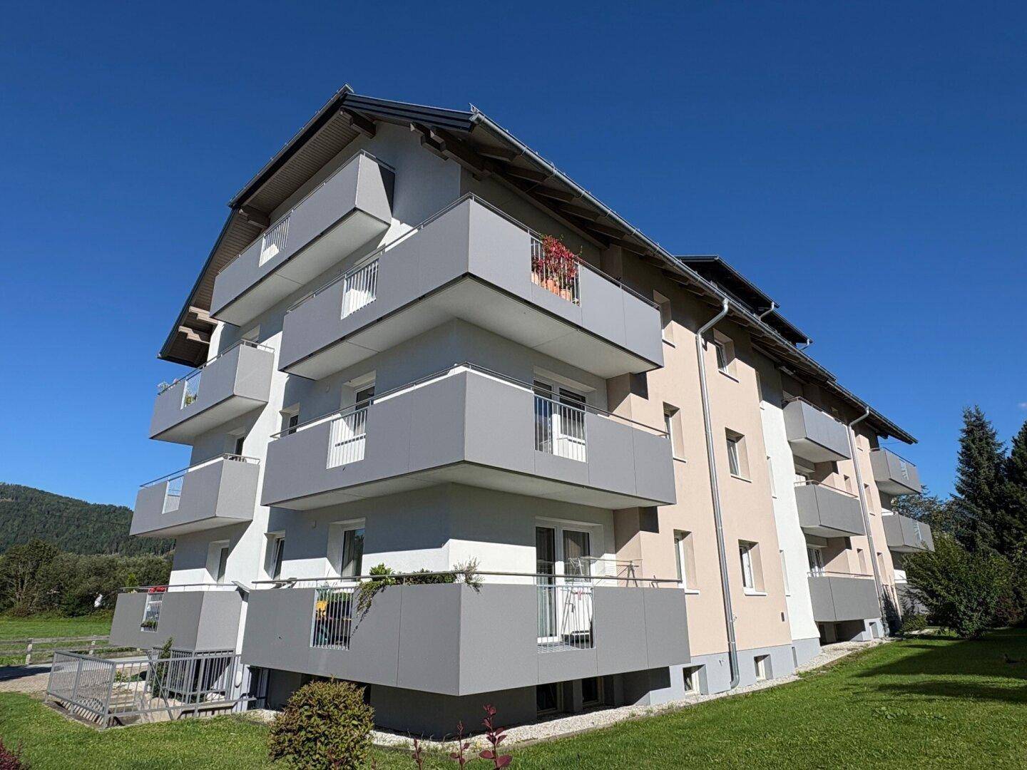 Seitenansicht des modernen Wohnhauses mit Balkonen, umgeben von grüner Natur und Bergen im Hintergrund.