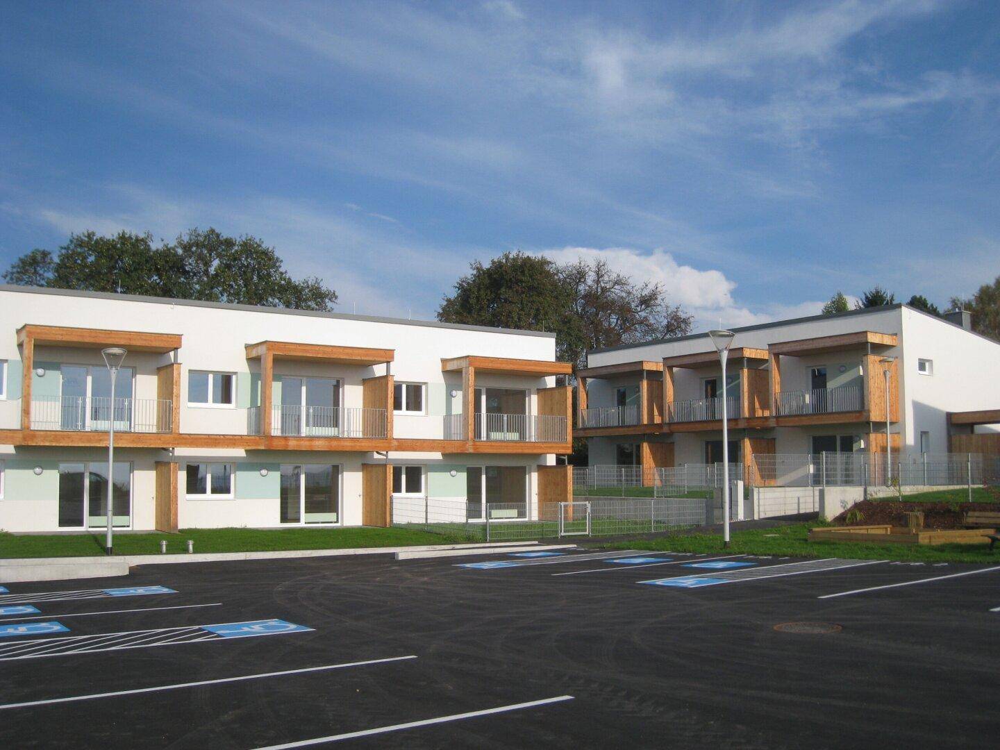 Zwei moderne Neubauten mit Balkonen und einem Parkplatz unter blauem Himmel.