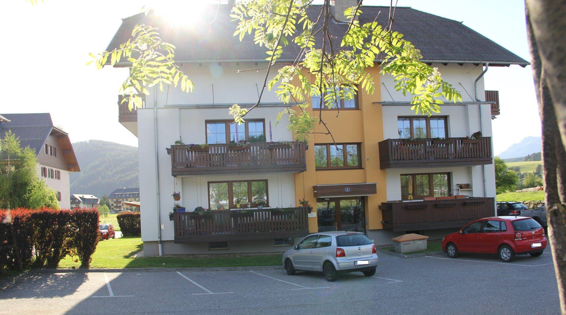 Frontansicht des Wohnhauses mit Balkonen und Parkplätzen, beleuchtet von der Abendsonne.