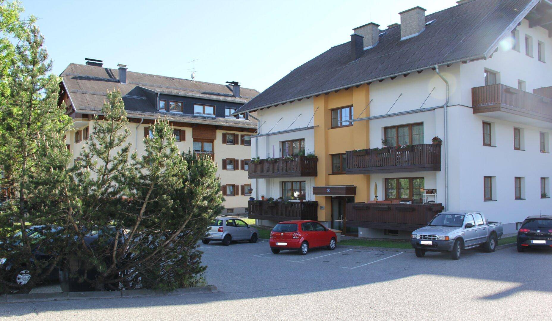 Mehrfamilienhaus mit Balkonen und Parkplätzen, umgeben von Bäumen und ländlicher Landschaft.