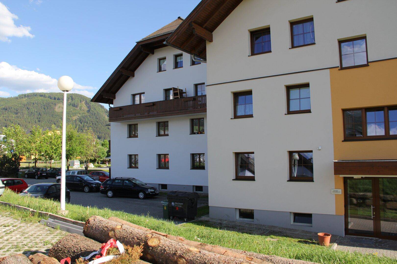 Seitenansicht eines Mehrfamilienhauses mit Holzbalkonen und Blick auf bewaldete Hügel.