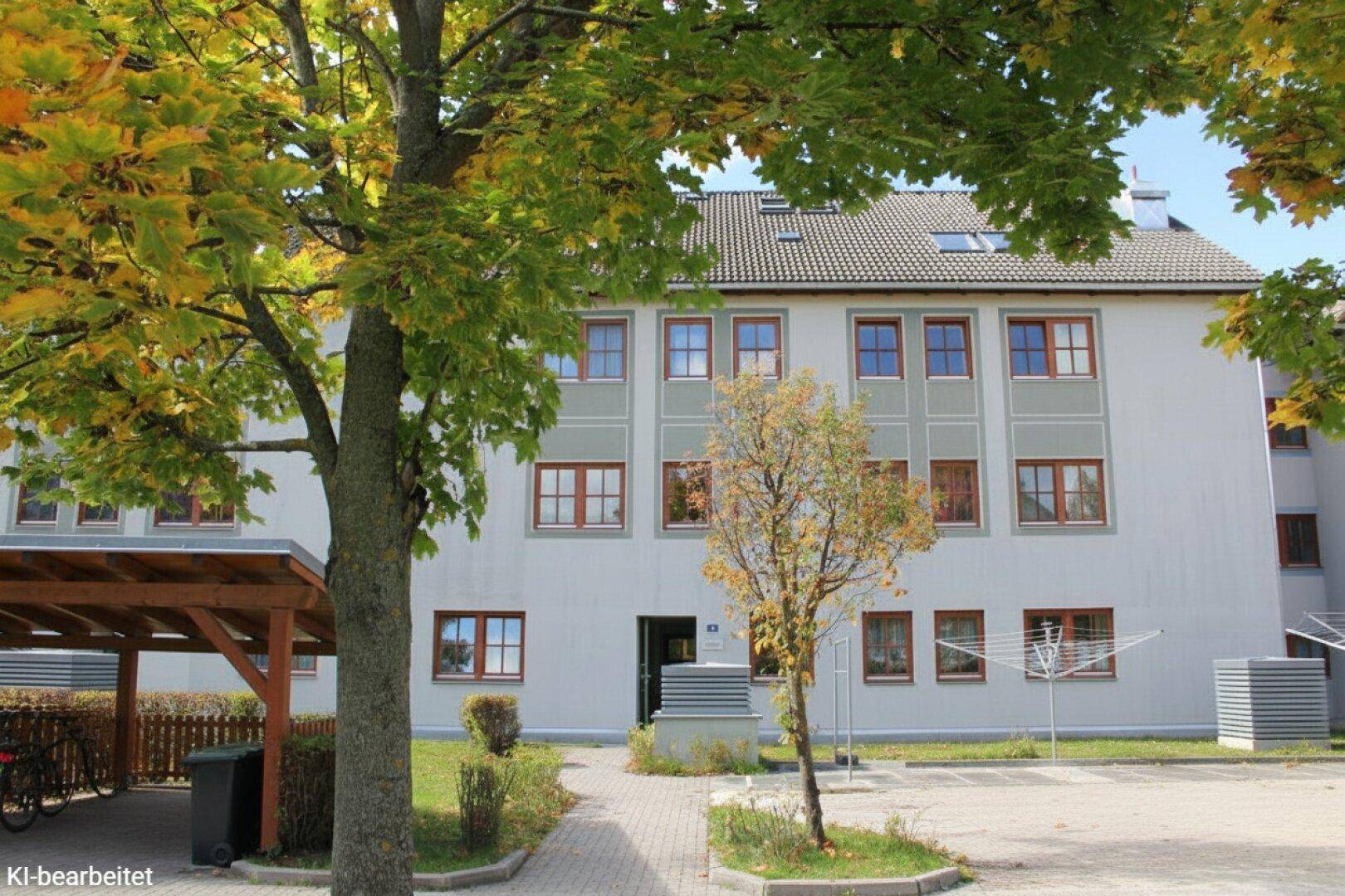 Rückansicht eines gepflegten Mehrfamilienhauses mit Carport und Bäumen im Vordergrund unter blauem Himmel.