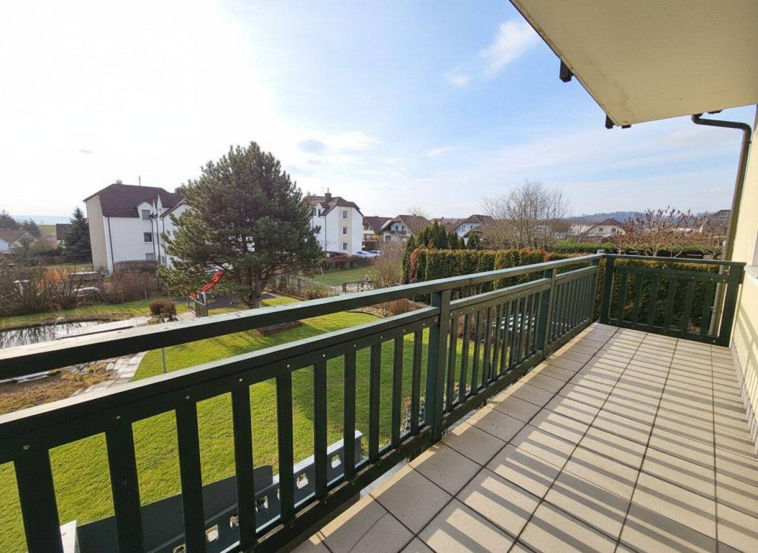 Sonniger Balkon mit grüner Brüstung und Blick auf den Gartenbereich und einen kleinen Teich.