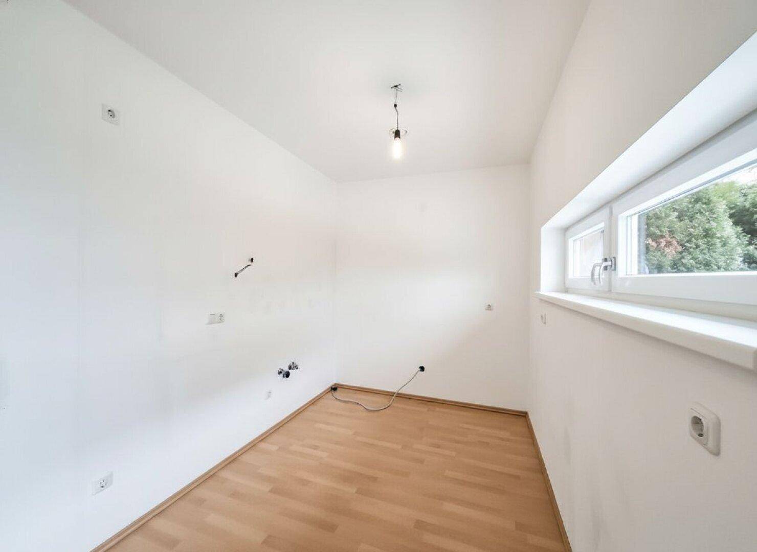 Leerer Raum mit Anschlüssen für eine Küche und einem Fenster mit Blick ins Grüne.