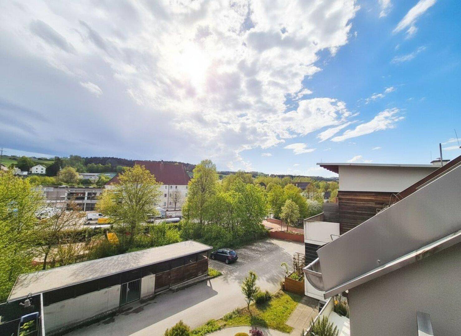 Weitläufiger Blick von einem Balkon auf die grüne Umgebung und die Dächer benachbarter Gebäude.