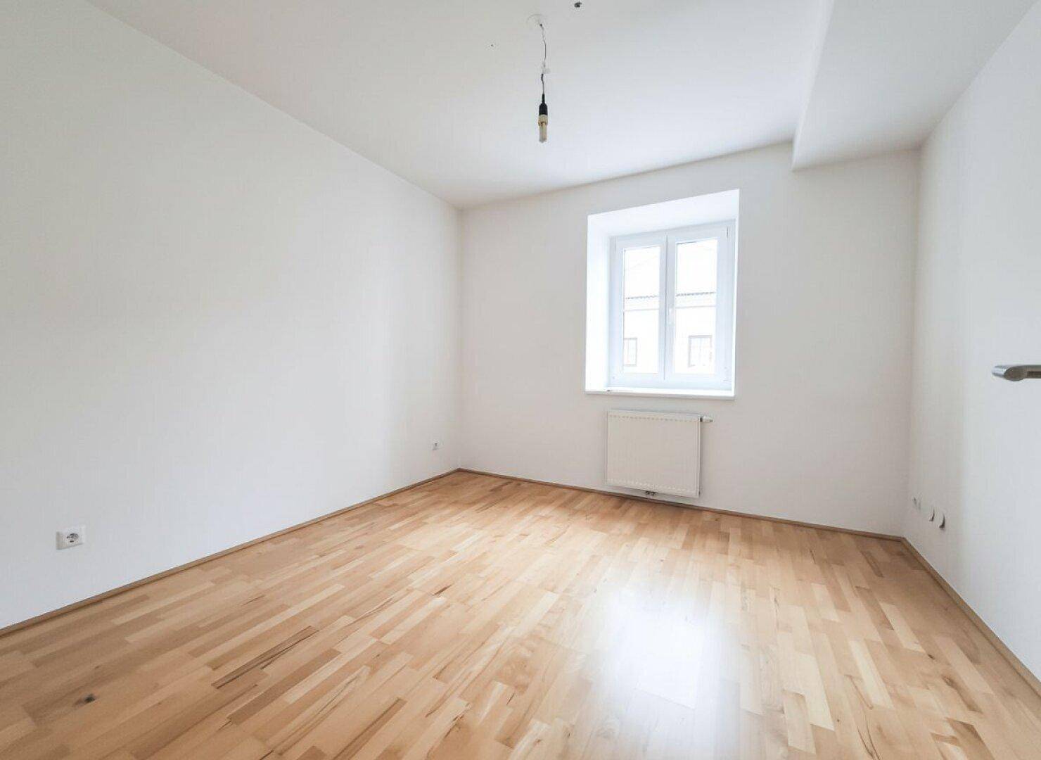 Helles Schlafzimmer mit Parkettboden und einem Fenster, das viel Tageslicht in den Raum bringt.