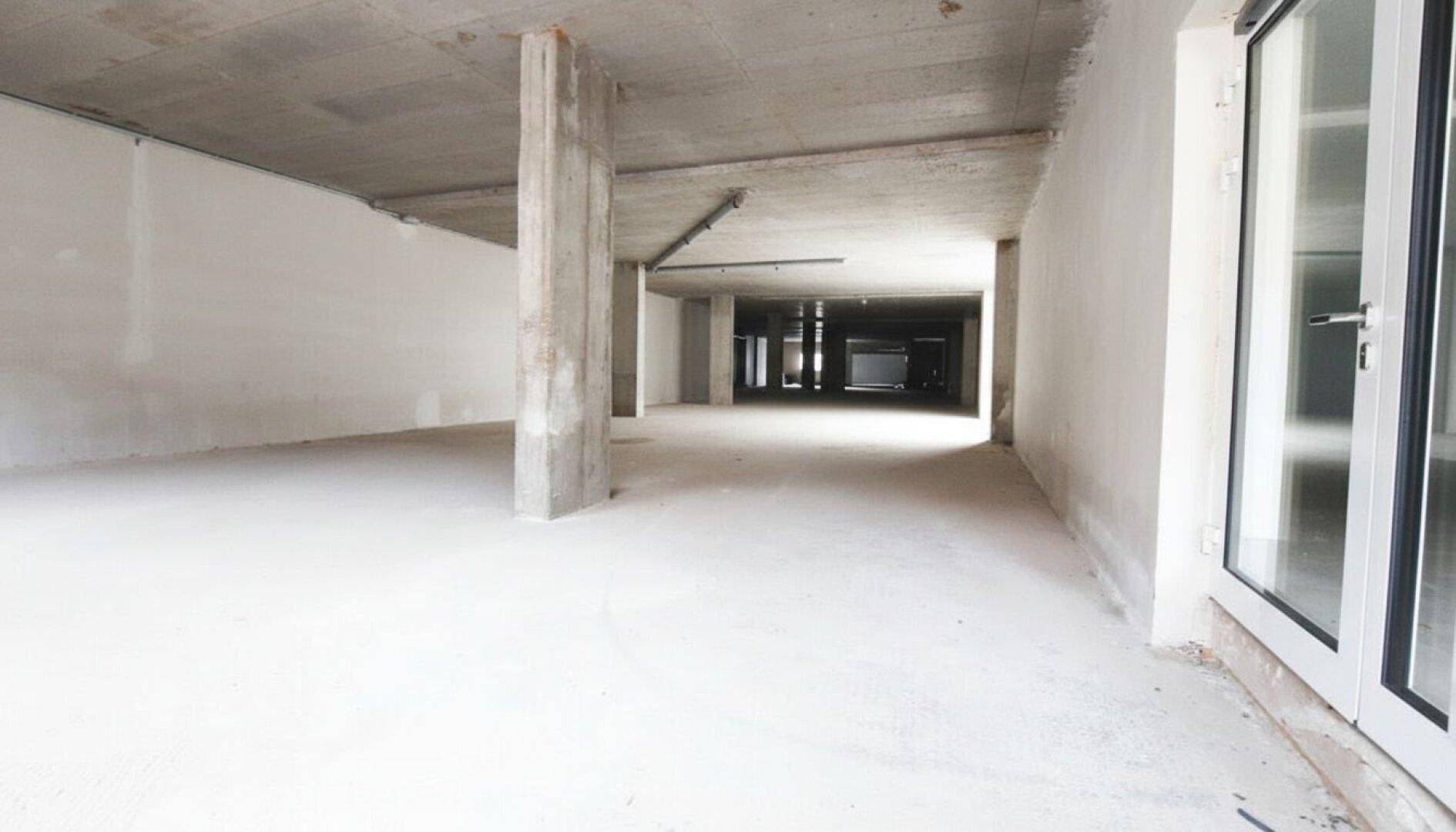 Weiter Blick in einen weitläufigen Gewerberaum im Rohbau mit Betonstütze und großen Fenstern.