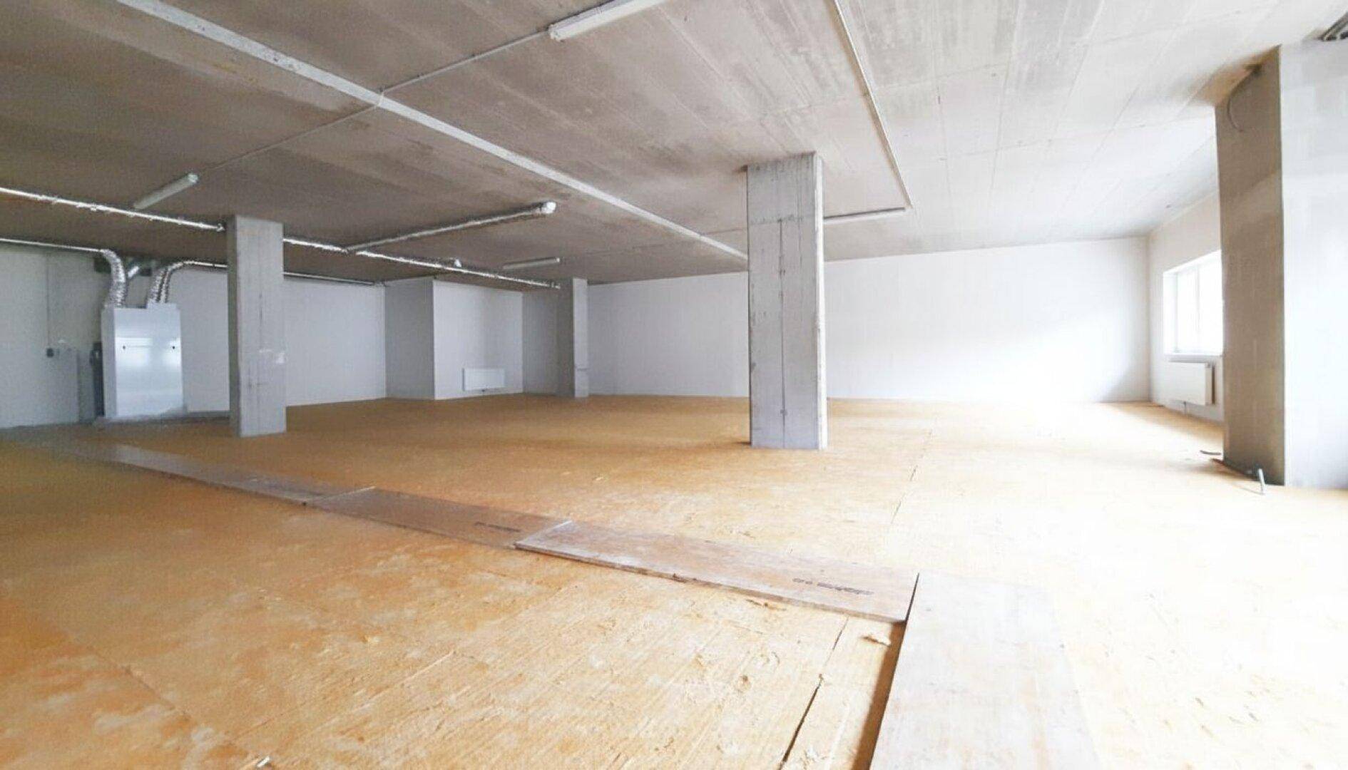 Weitläufiger Innenraum im Rohbau mit Betonstützen, hohen Decken und OSB-Platten auf dem Boden.