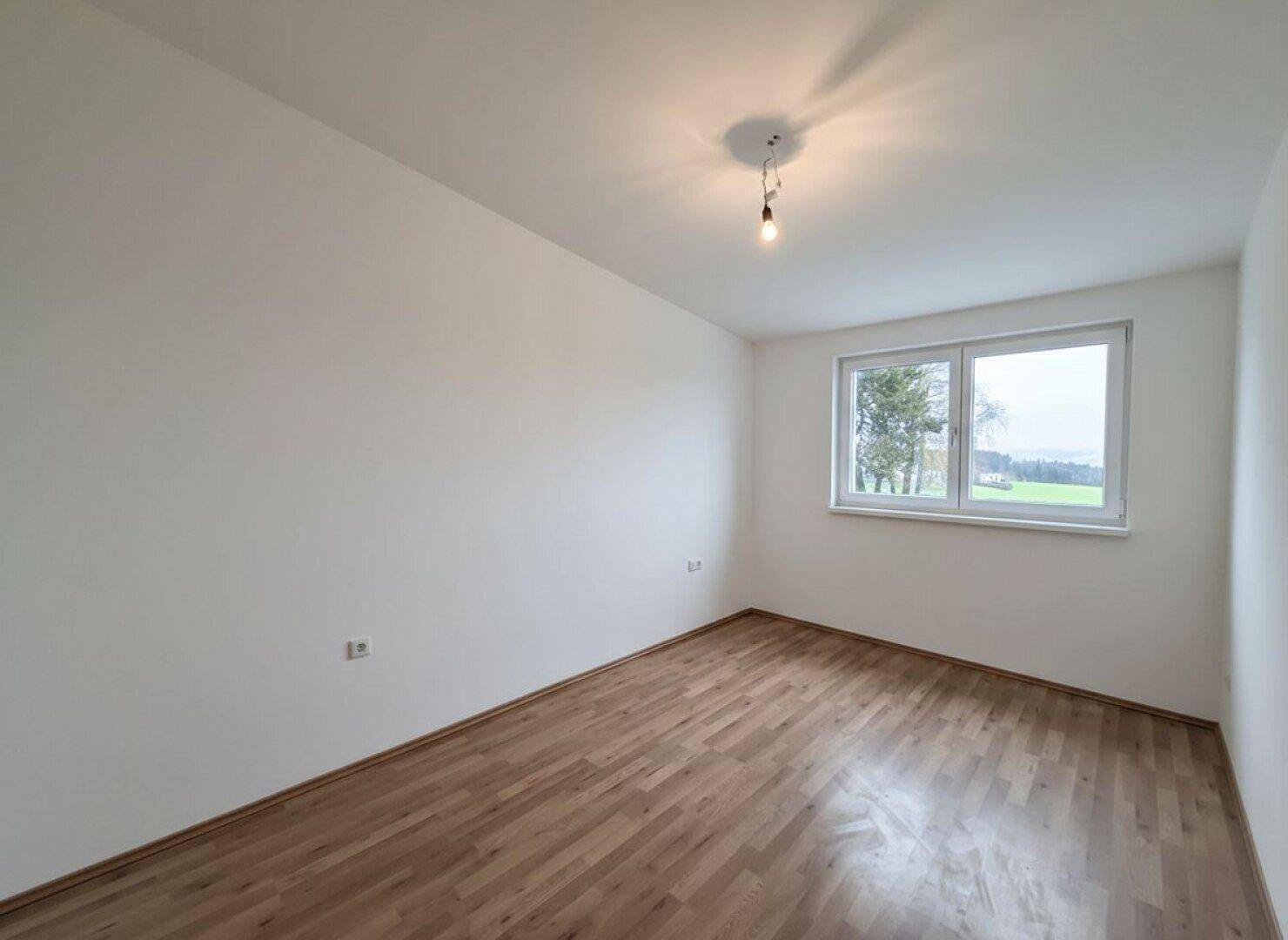 Helles Schlafzimmer mit Parkettboden und großem Fenster, das einen grünen Ausblick bietet.