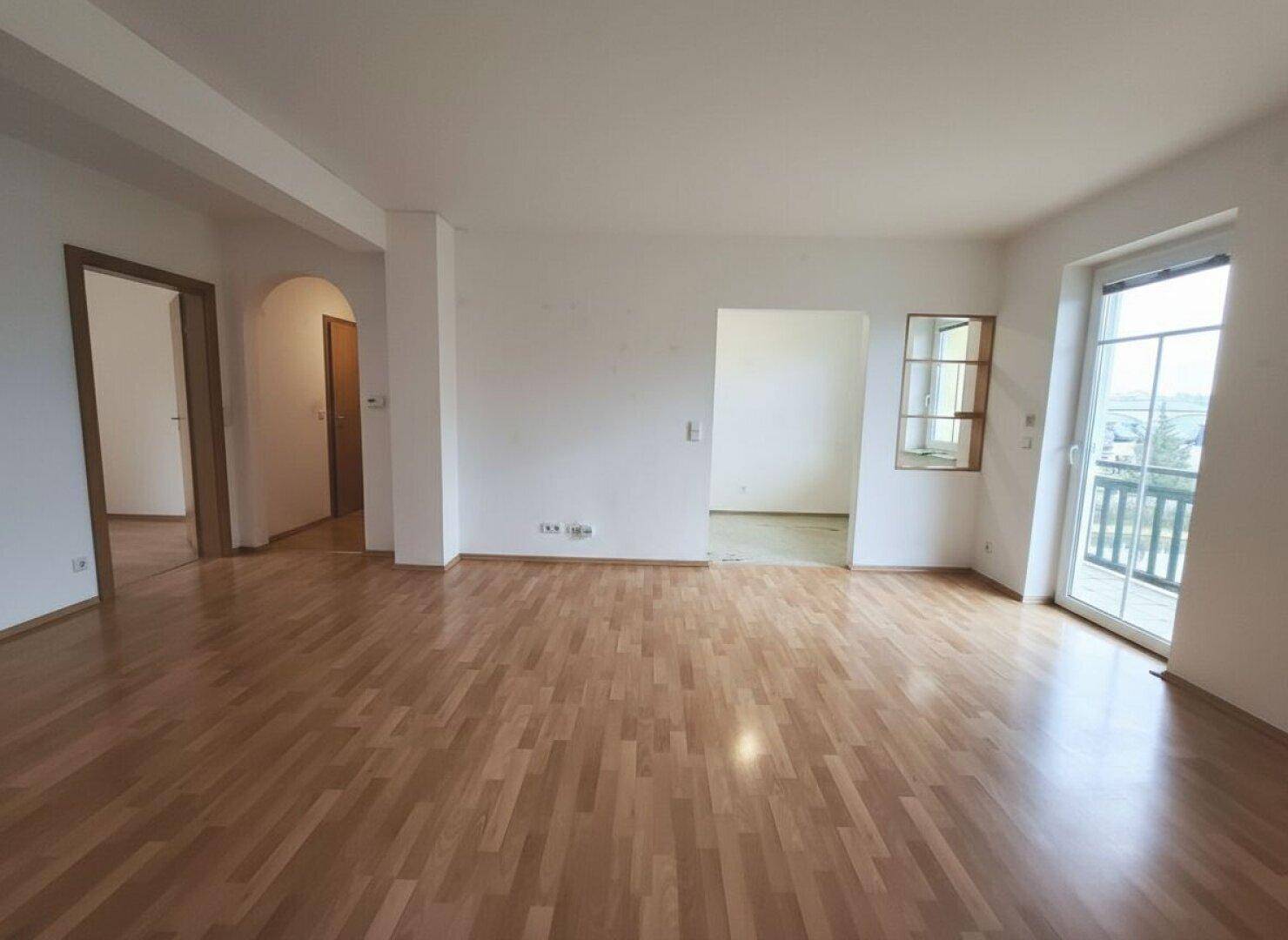 Helles Wohnzimmer mit Parkettboden, das zu weiteren Räumen und einem Balkon führt und viel Licht bietet.