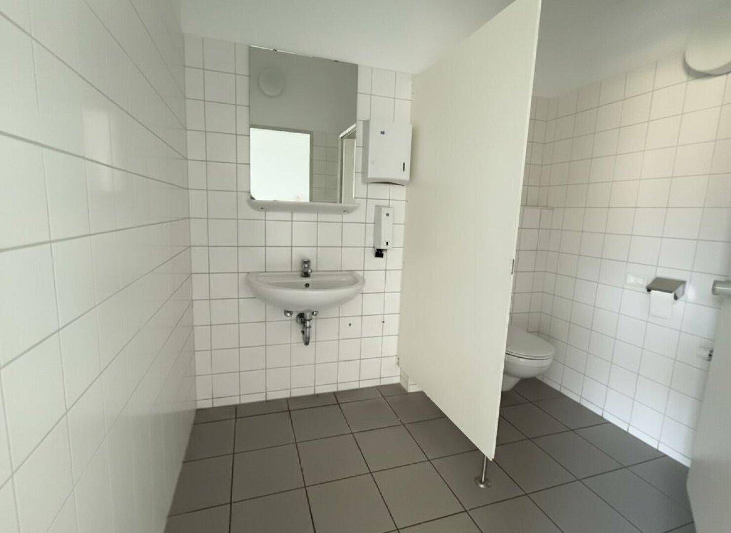Badezimmer mit Waschbecken, Toilette und weißen Wandfliesen, getrennt durch eine Trennwand.