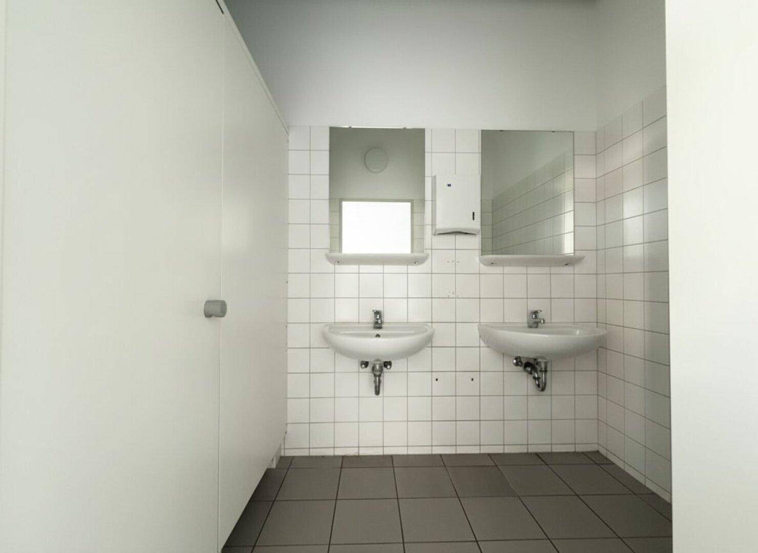 Sanitärbereich mit zwei Waschbecken, weißen Fliesen an den Wänden und grauem Fliesenboden.
