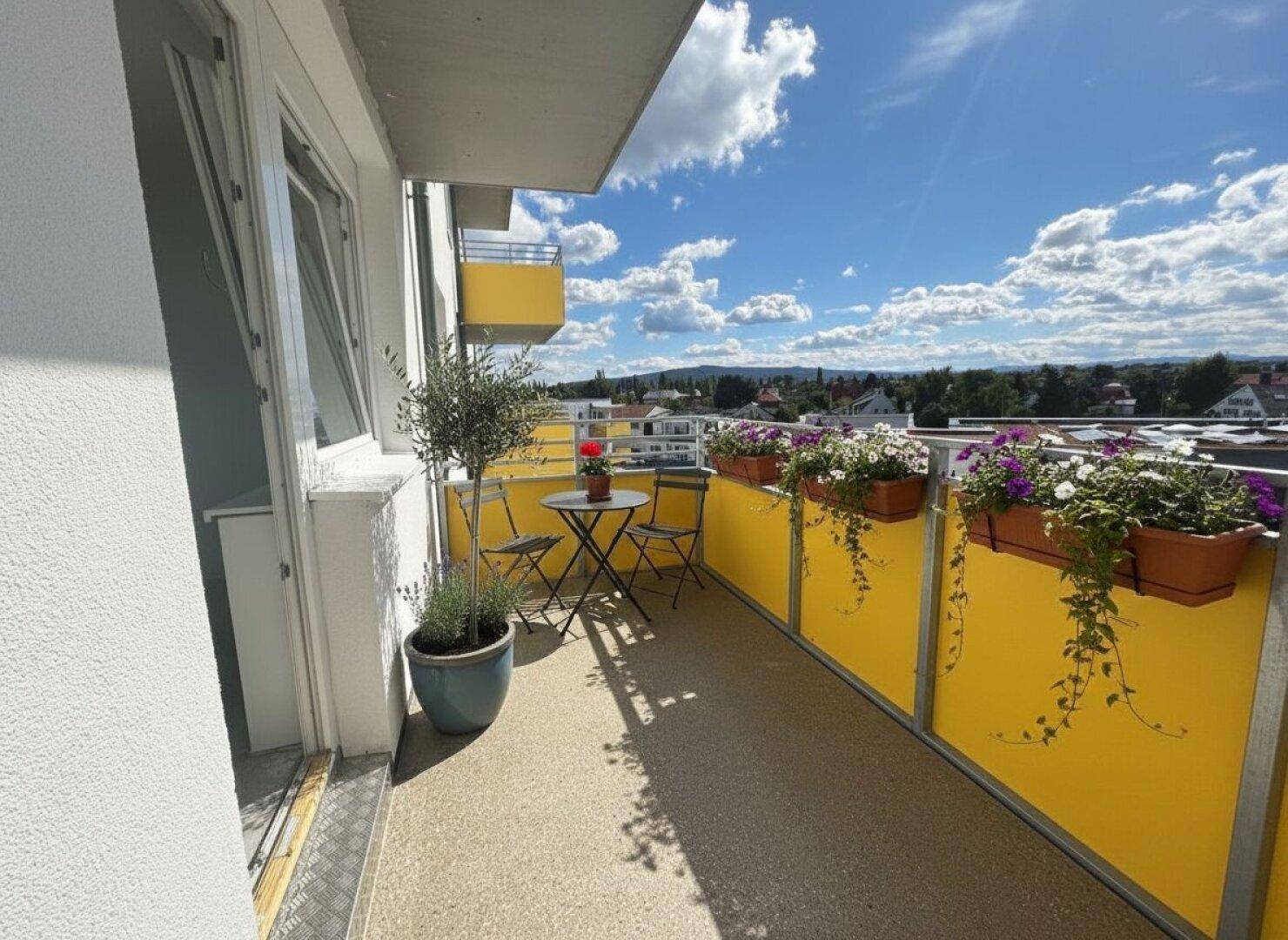 Langer Balkon mit gelben Sichtschutzwänden, Blumenkästen und Blick auf die Umgebung.