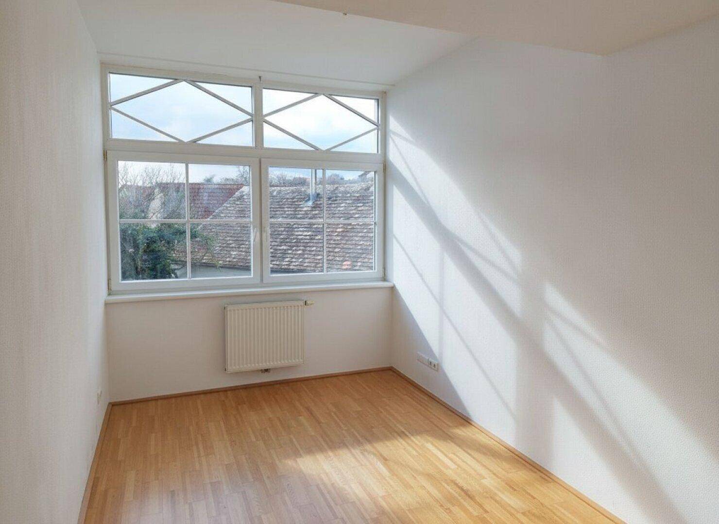Helles Zimmer mit Parkettboden und großem Fenster mit Blick auf die Dächer.