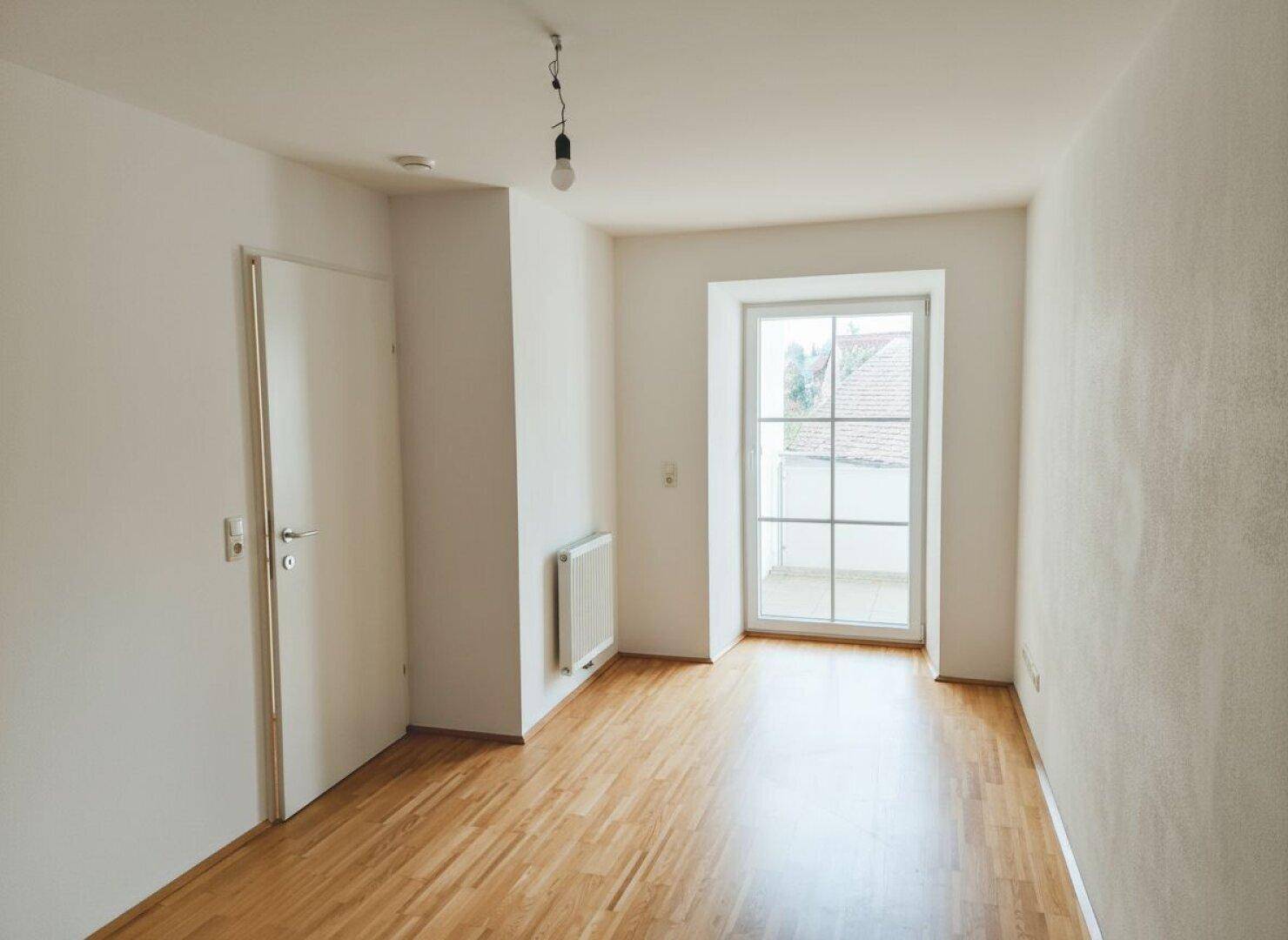 Leeres Zimmer mit Parkettboden, weißen Wänden und Zugang zu einem französischen Balkon.