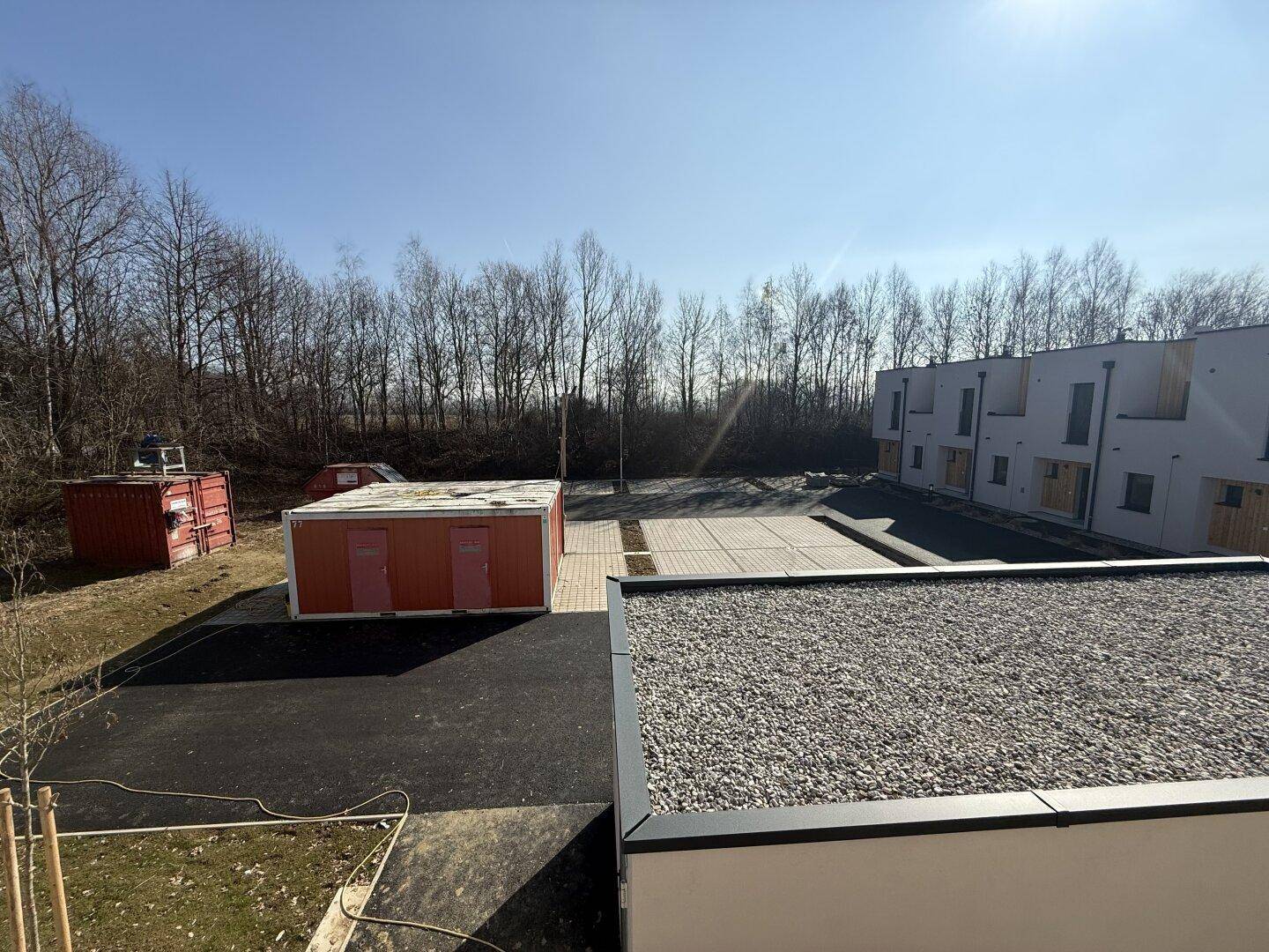 Außenansicht mit Flachdach, Parkplätzen und weiteren Neubauten im Hintergrund unter blauem Himmel.
