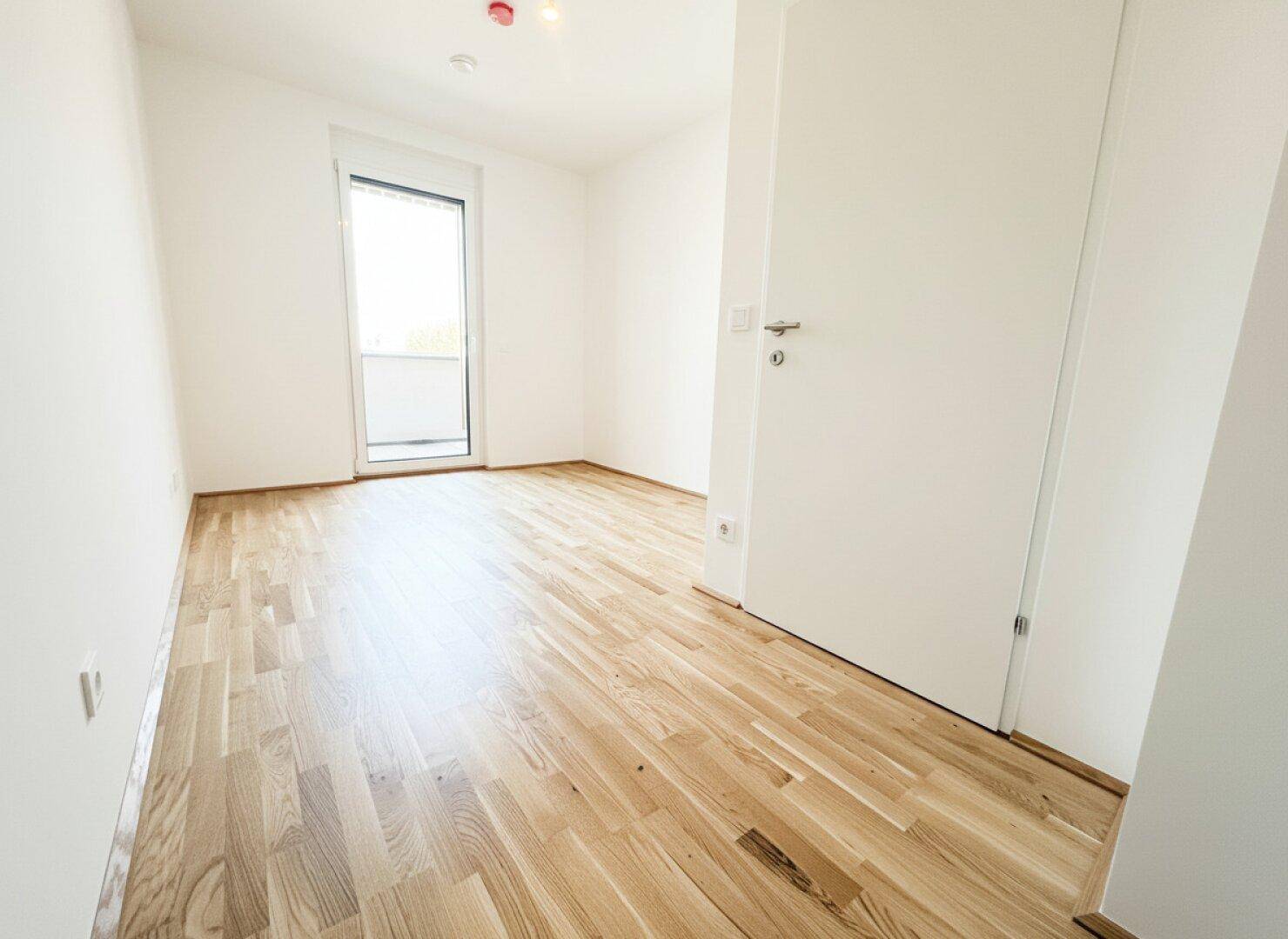 Unmöbliertes Zimmer mit Parkettboden und einem bodentiefen Fenster, das viel Licht hereinlässt.