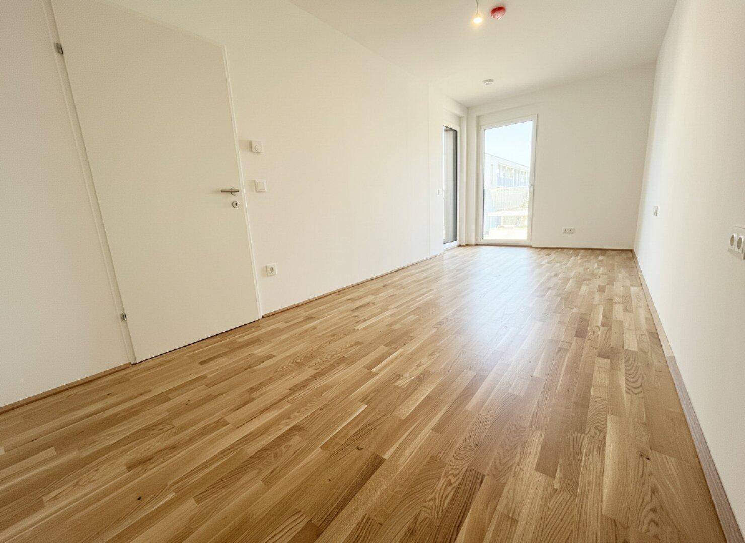 Helles Zimmer mit Parkettboden und Zugang zu einem Außenbereich durch eine große Glastür.