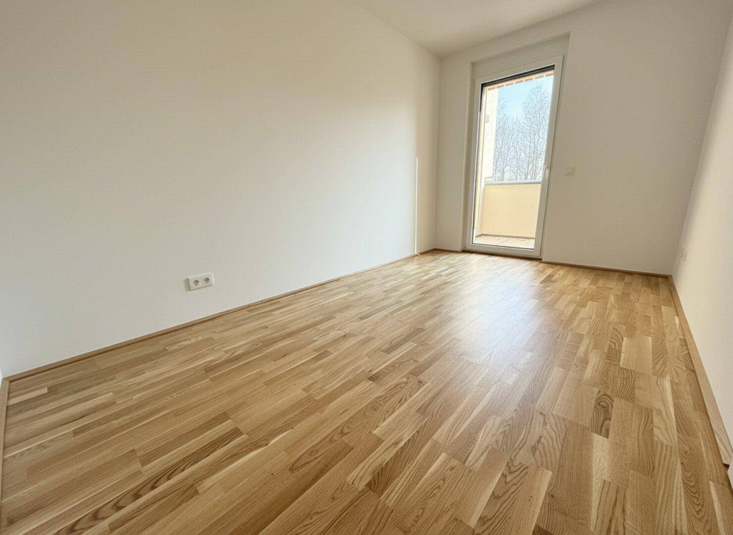 Leeres Zimmer mit Parkettboden und einem bodentiefen Fenster, das einen französischen Balkon andeutet.