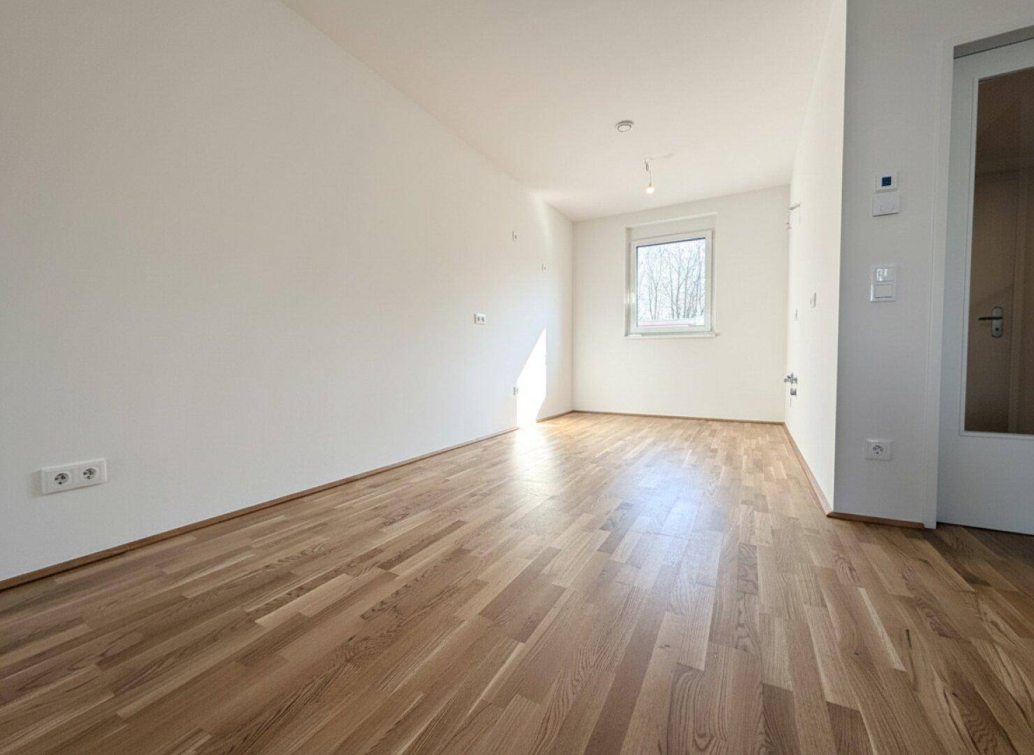 Helles und unmöbliertes Zimmer mit Parkettboden und einem Fenster mit Blick ins Grüne.
