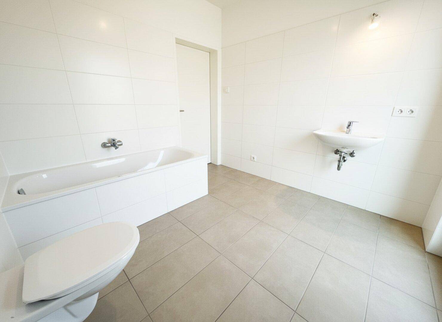 Modernes Badezimmer mit Badewanne, WC und Waschbecken, komplett gefliest in Weiß und Grau.