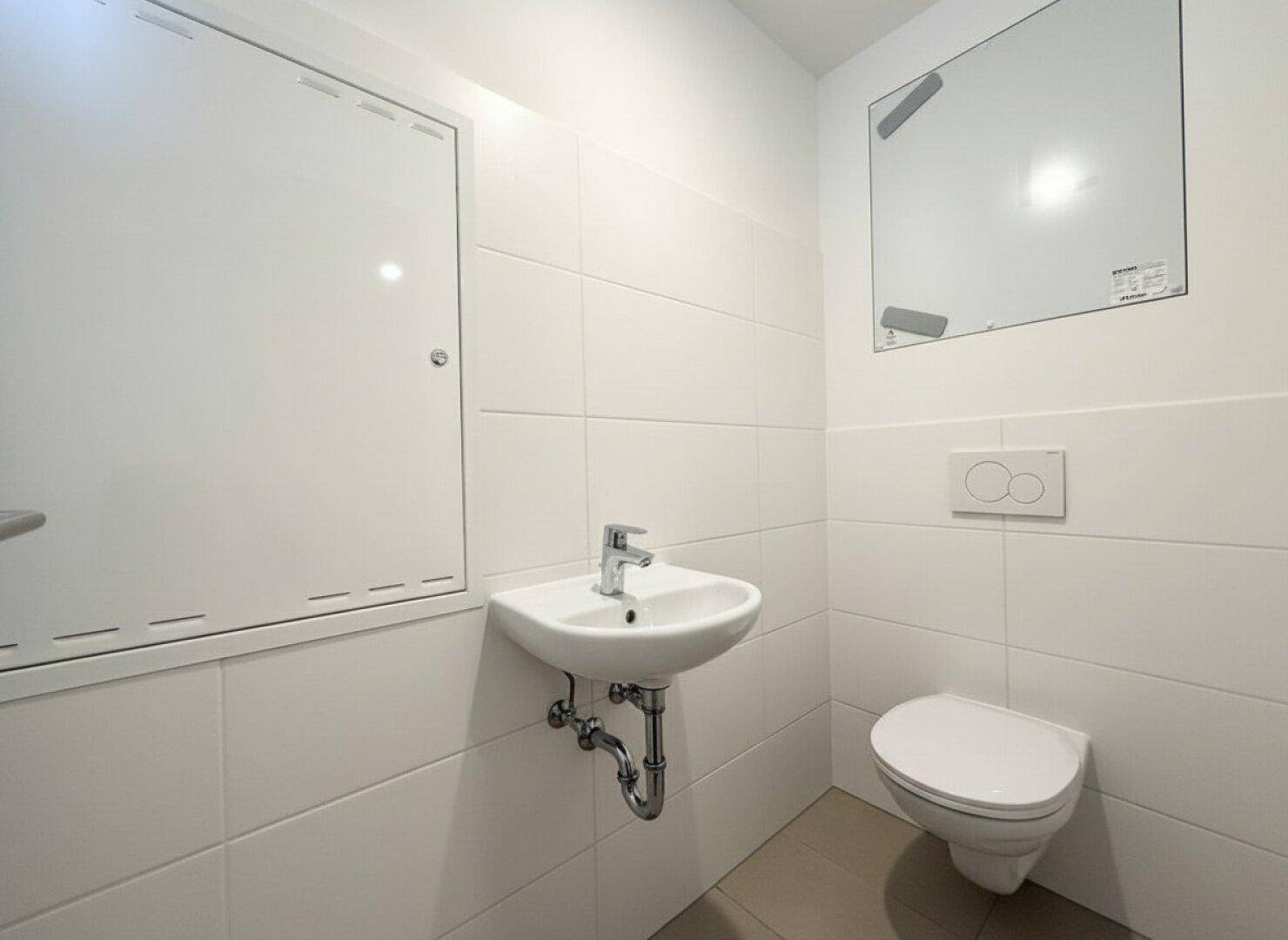 Gäste-WC mit Wand-WC und kleinem Waschbecken, vollständig gefliest in Weiß und Beige.