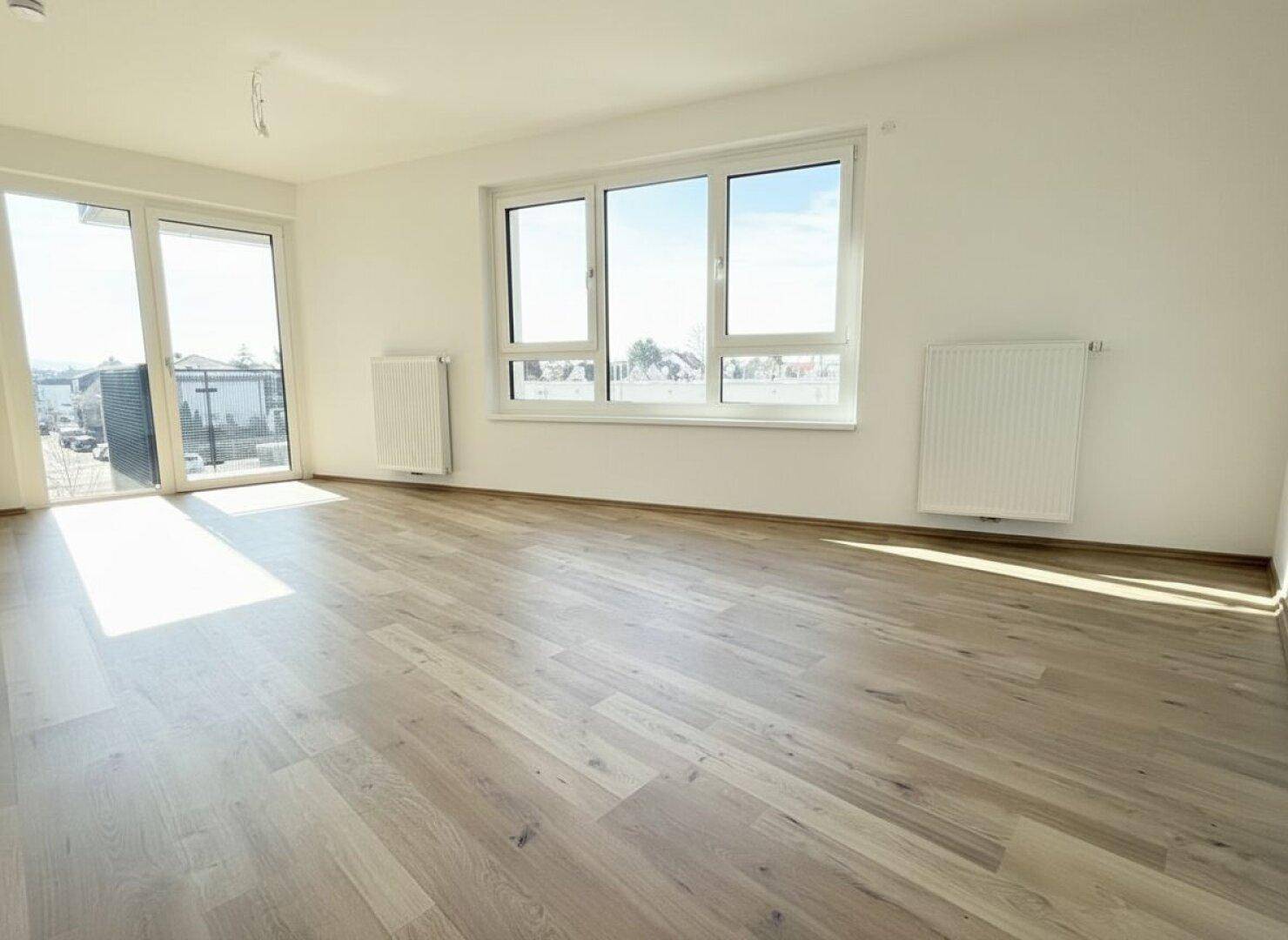 Lichtdurchfluteter Wohnraum mit hellem Holzboden und großen Fenstern, die viel Tageslicht hereinlassen.