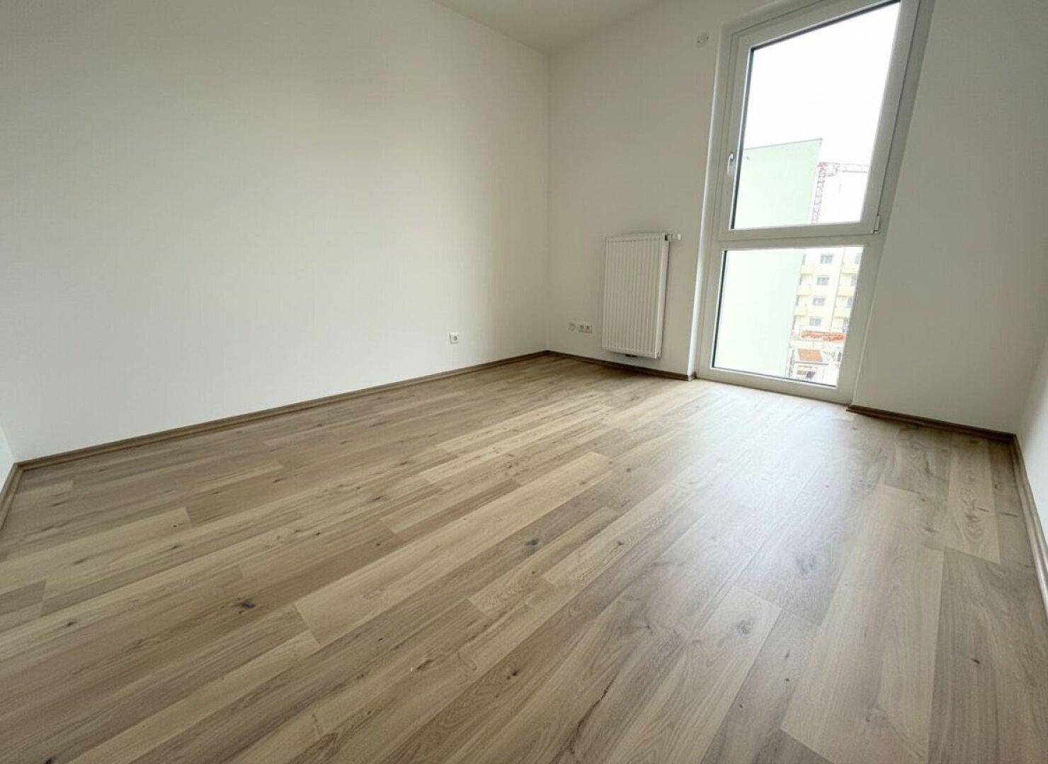 Modernes Schlafzimmer mit hellem Laminatboden und einem hohen Fenster, das viel Licht hereinlässt.