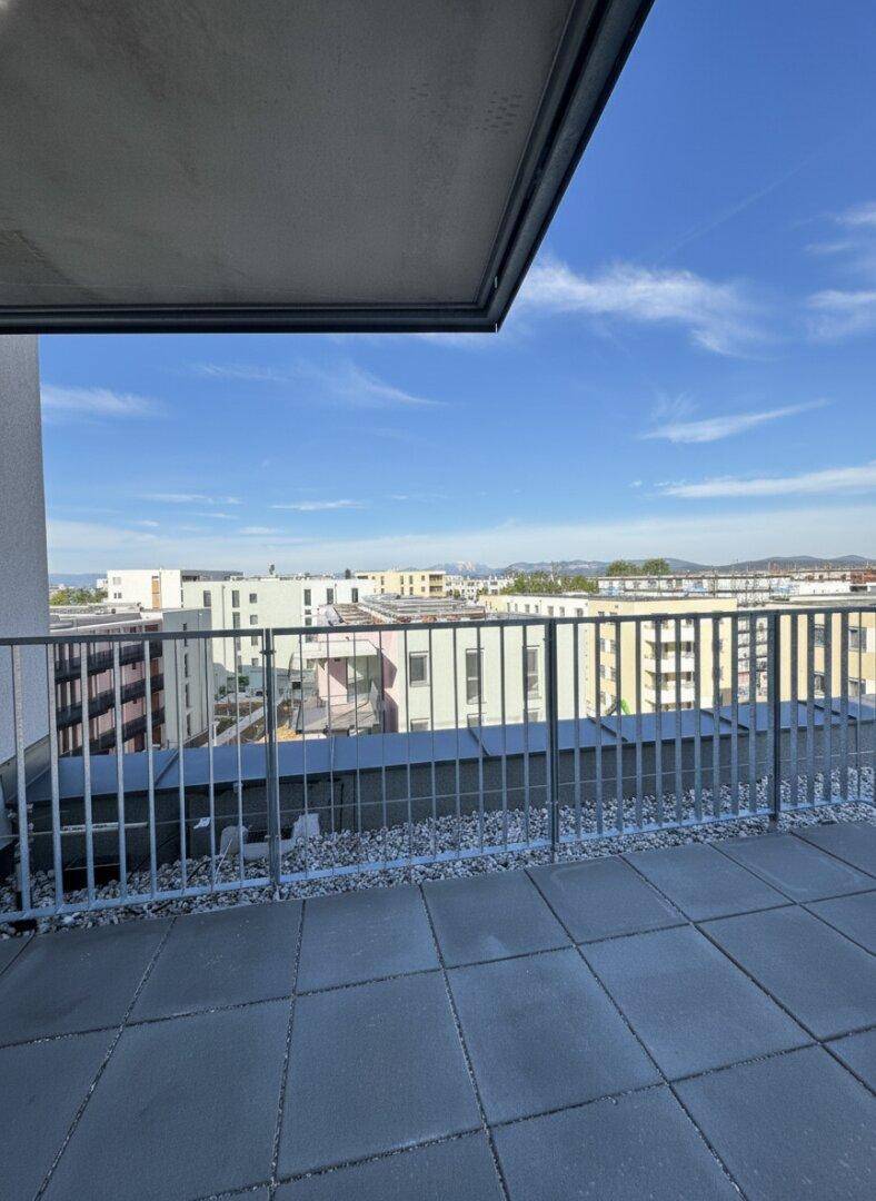 Blick von einem modernen Balkon mit Fliesenboden über die Stadtlandschaft und entfernte Berge unter blauem Himmel.