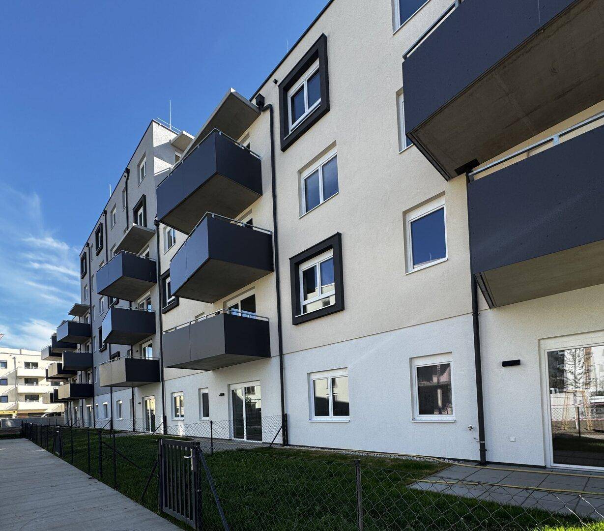 Moderne Wohngebäude mit weißer Fassade, dunklen Balkonen und gepflegten Grünflächen unter blauem Himmel.