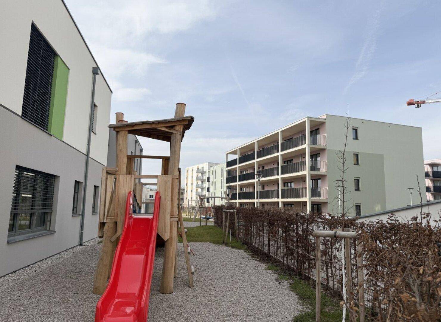 Spielplatz mit roter Rutsche und Holzstruktur zwischen modernen Wohngebäuden mit Balkonen.