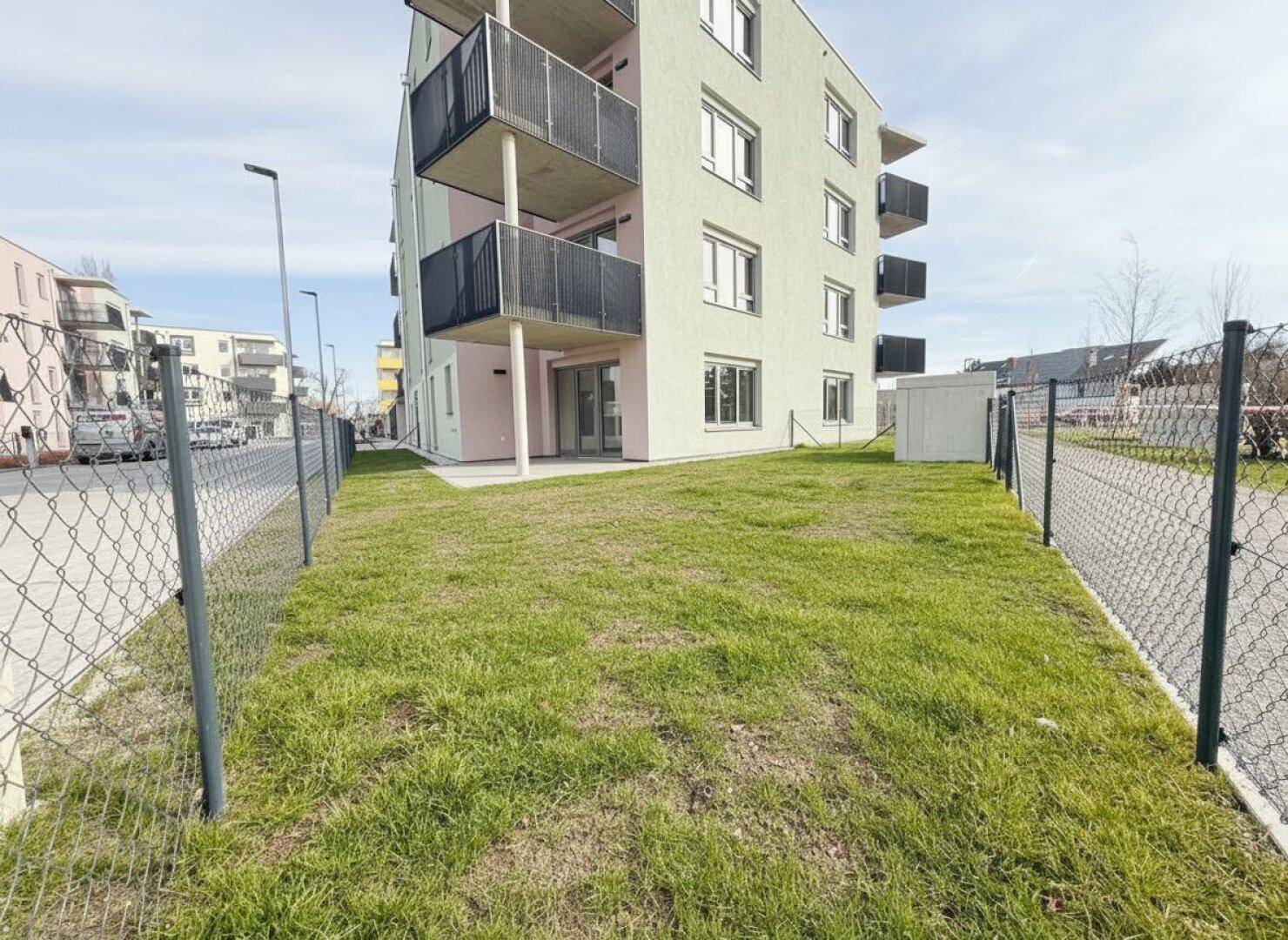 Neubauwohnung mit Balkonen und einer kleinen Rasenfläche, umzäunt von einem Maschendrahtzaun.